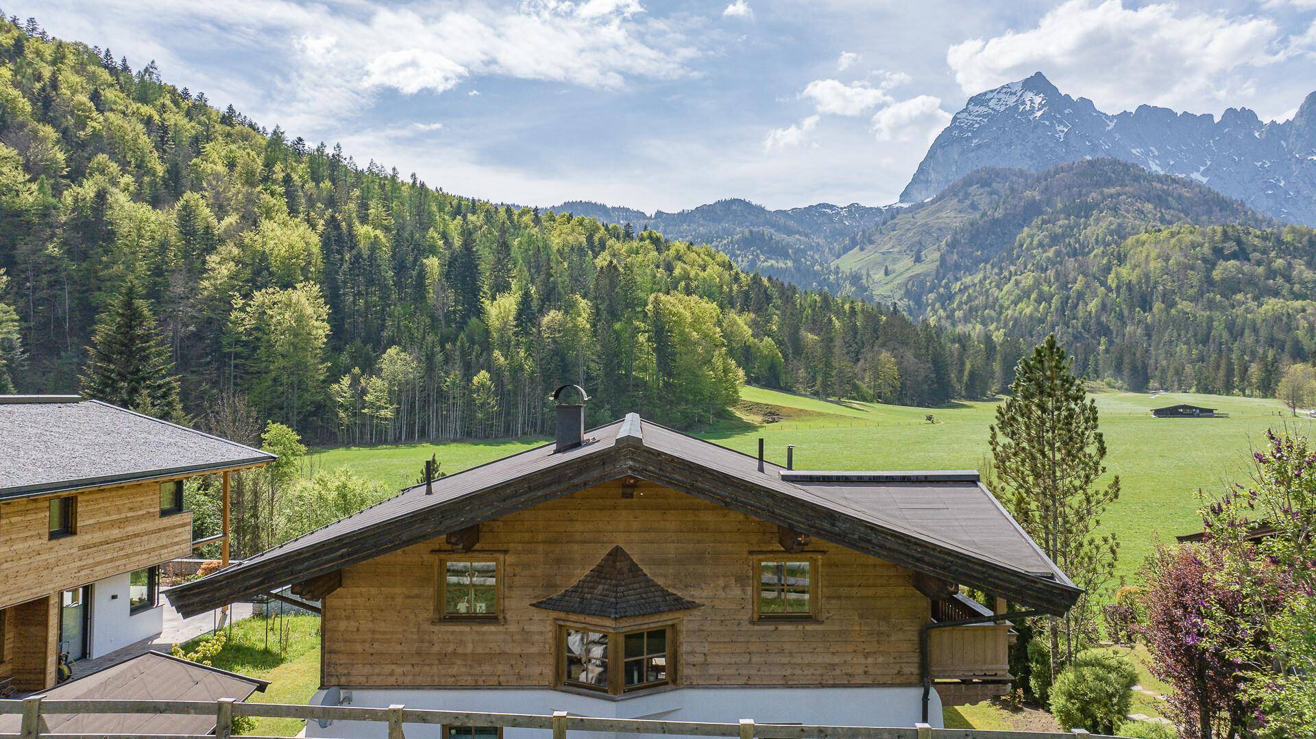 Rückansicht des Holzhauses mit Satteldach und weitem Blick auf grüne Wiesen und Berge.