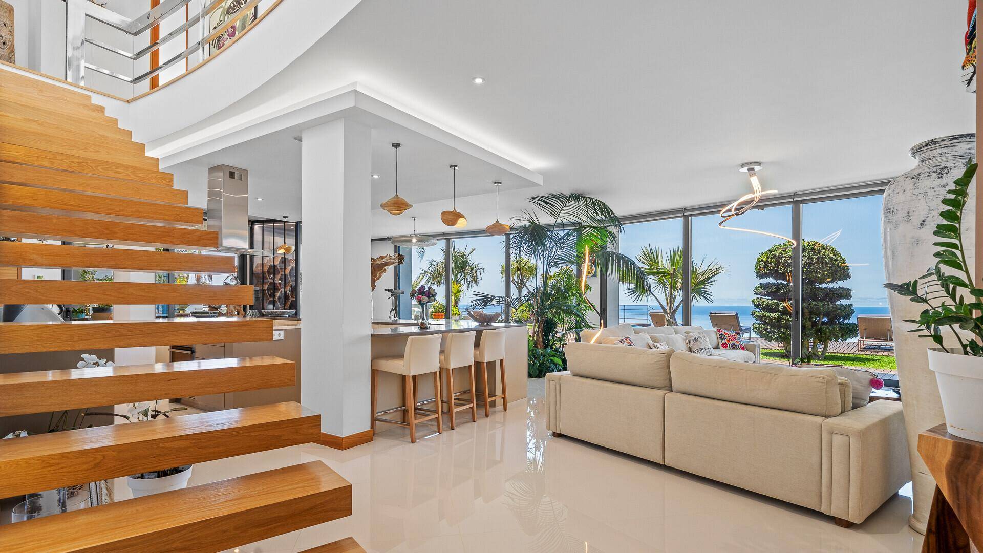 Offener Wohnbereich mit eleganter Holztreppe, bequemen Sofas und Meerblick durch große Panoramafenster.