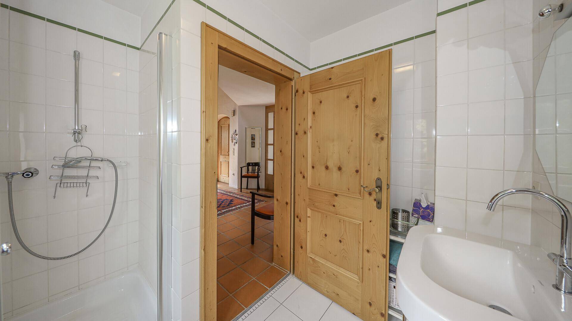 Badezimmer mit weißen Fliesen, Duschbereich und Holztür, die zum Flur führt.