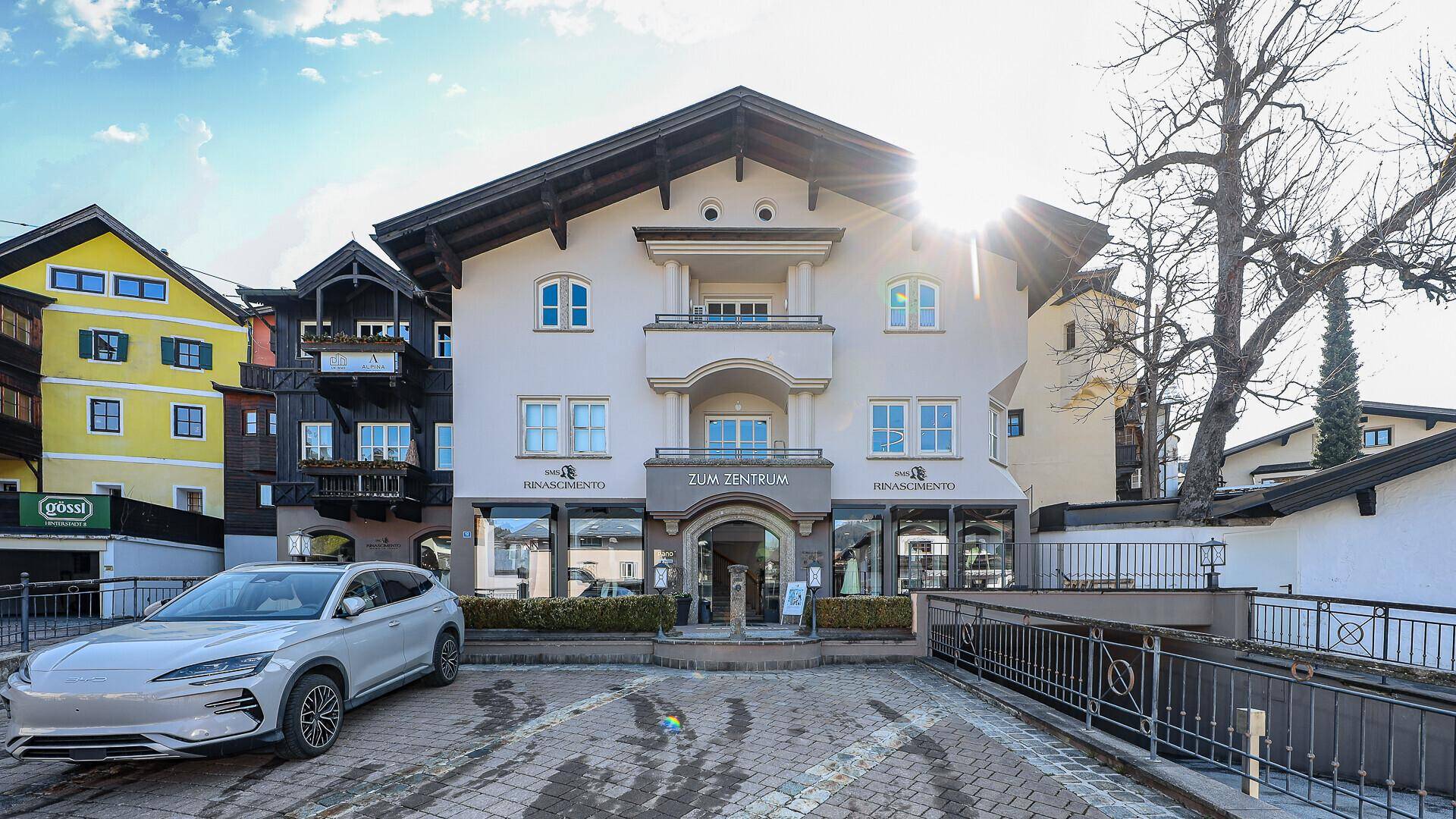 Frontansicht eines Gebäudes im alpinen Stil mit Parkplatz und einem Auto davor.
