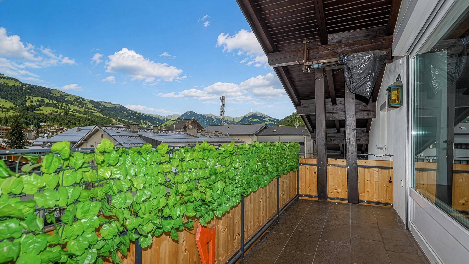 Langer Balkon mit Holzboden und Bergblick, geschützt durch eine grüne Hecke.
