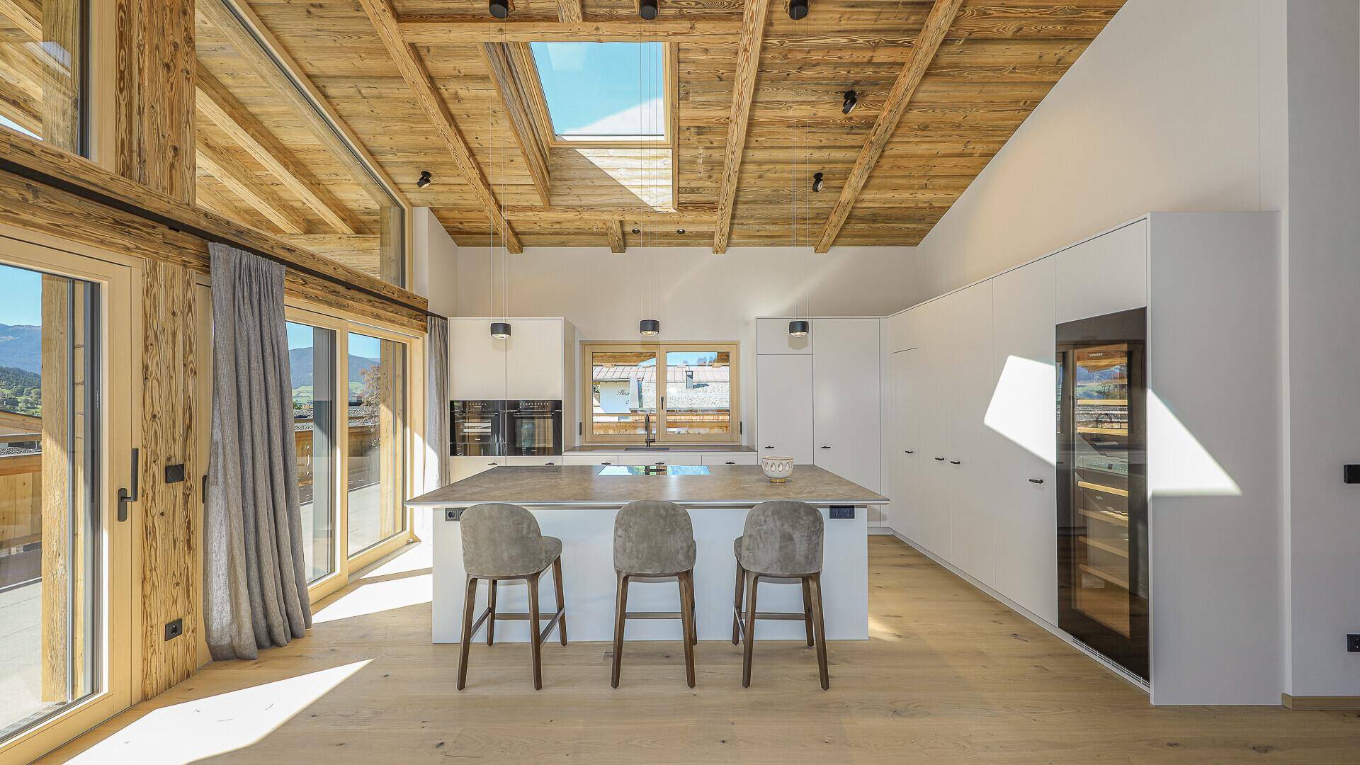 Moderne Einbauküche mit Kochinsel, Barhockern und integrierten Geräten unter Holzbalkendecke.