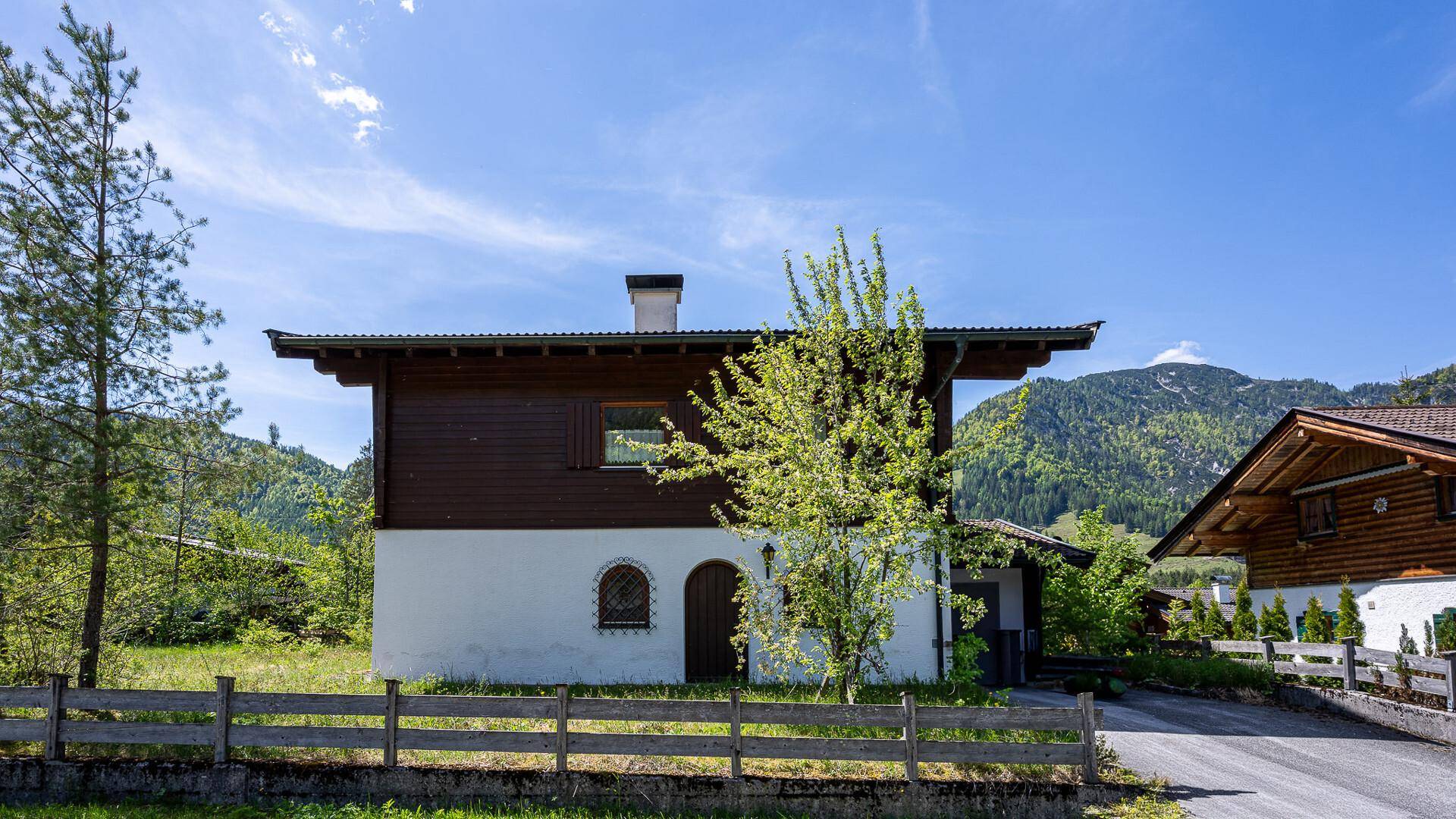 Frontansicht eines traditionellen Chalets mit dunkler Holzfassade und weißem Sockel vor Bergpanorama.