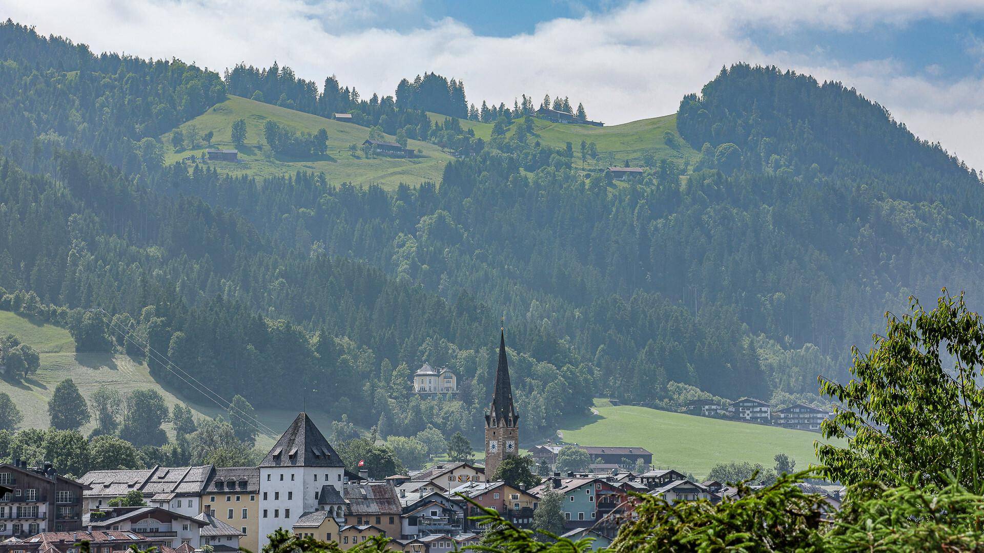 Panoramablick auf ein malerisches Bergdorf mit grünen Hügeln und bewaldeten Bergen im Hintergrund.
