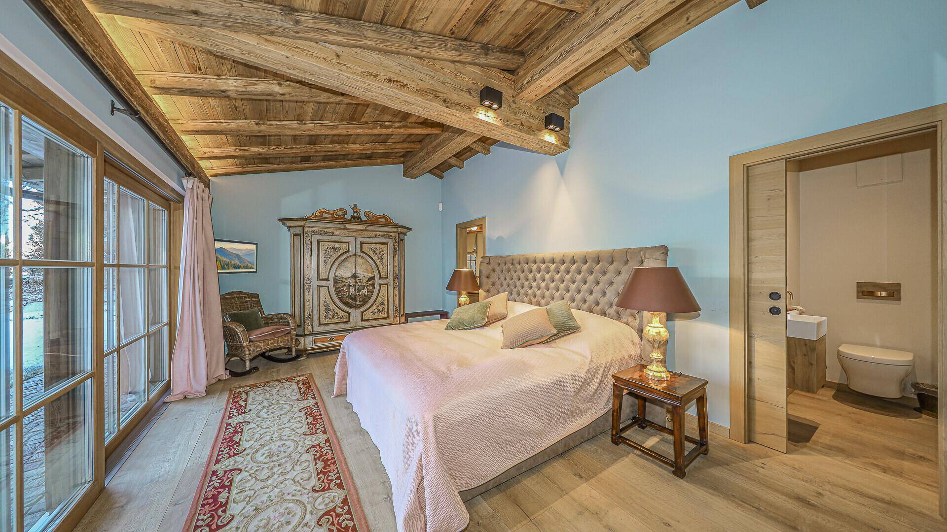 Schlafzimmer mit Holzbalkendecke, großem Bett und Zugang zu einem kleinen WC, im alpinen Stil.