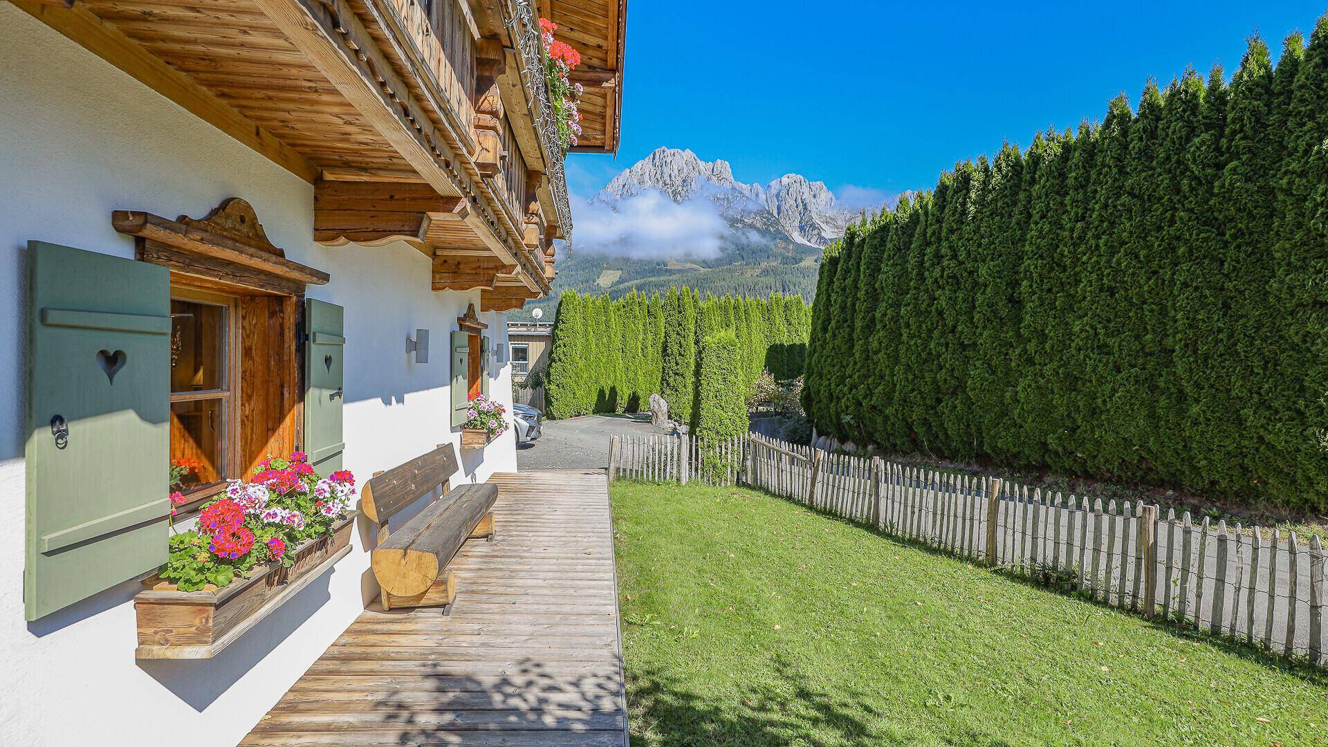 Traditionelles Haus mit Holzbalkonen, grüner Hecke und Bergblick unter blauem Himmel.