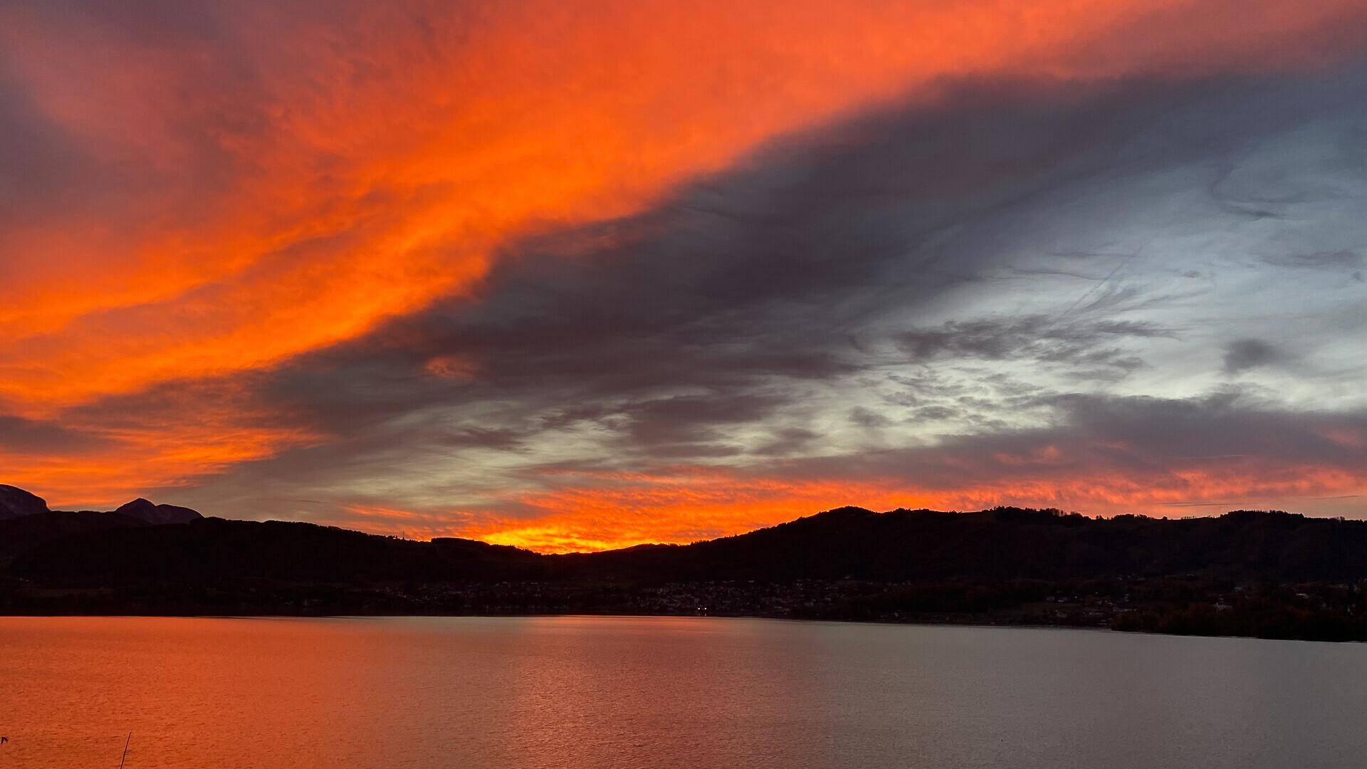 Dramatischer Sonnenuntergang über einem See mit leuchtend orangefarbenen und grauen Wolken am Horizont.
