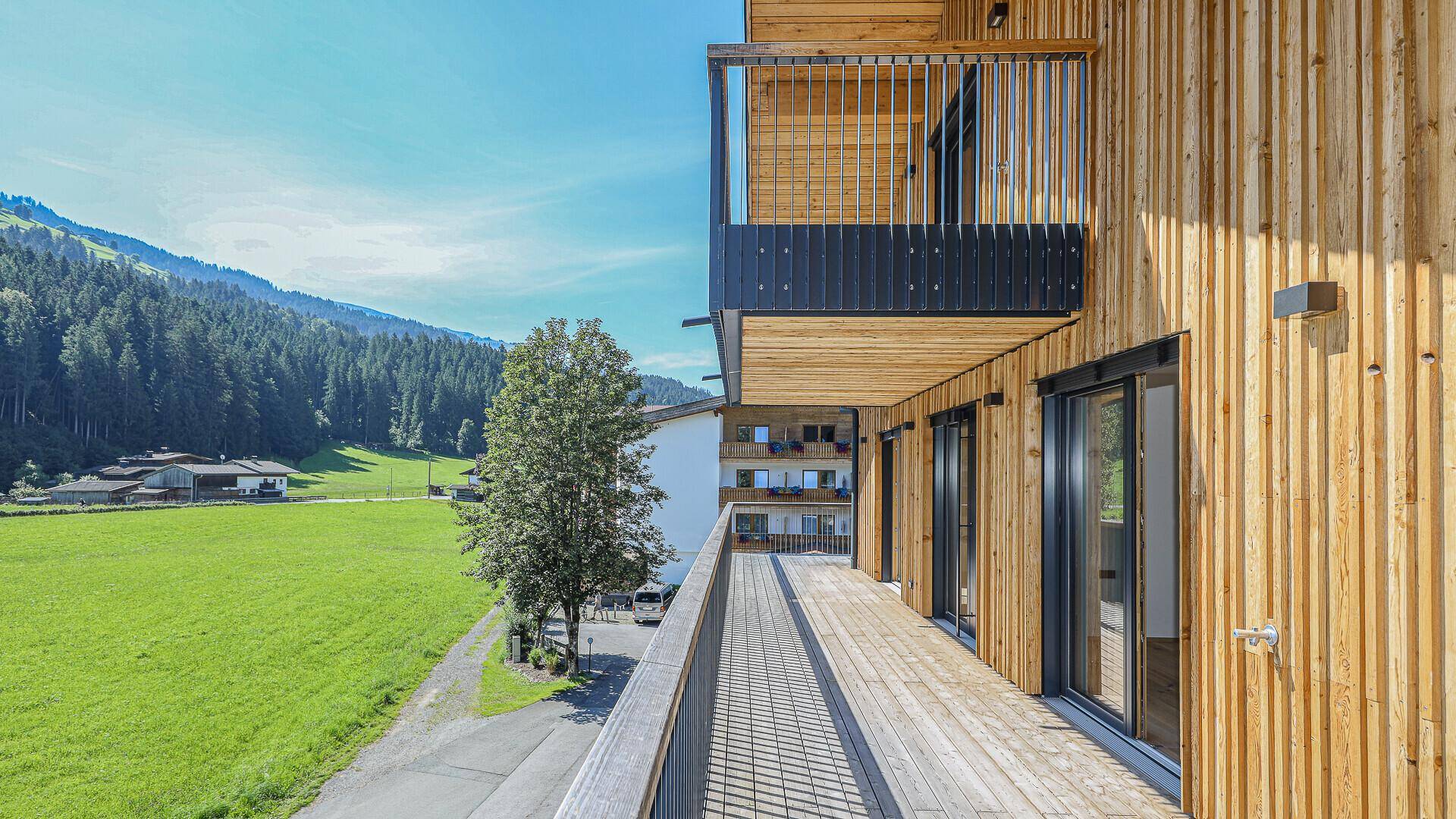 Breiter Holzbalkon mit Blick auf eine grüne Wiese und die bewaldeten Berge.