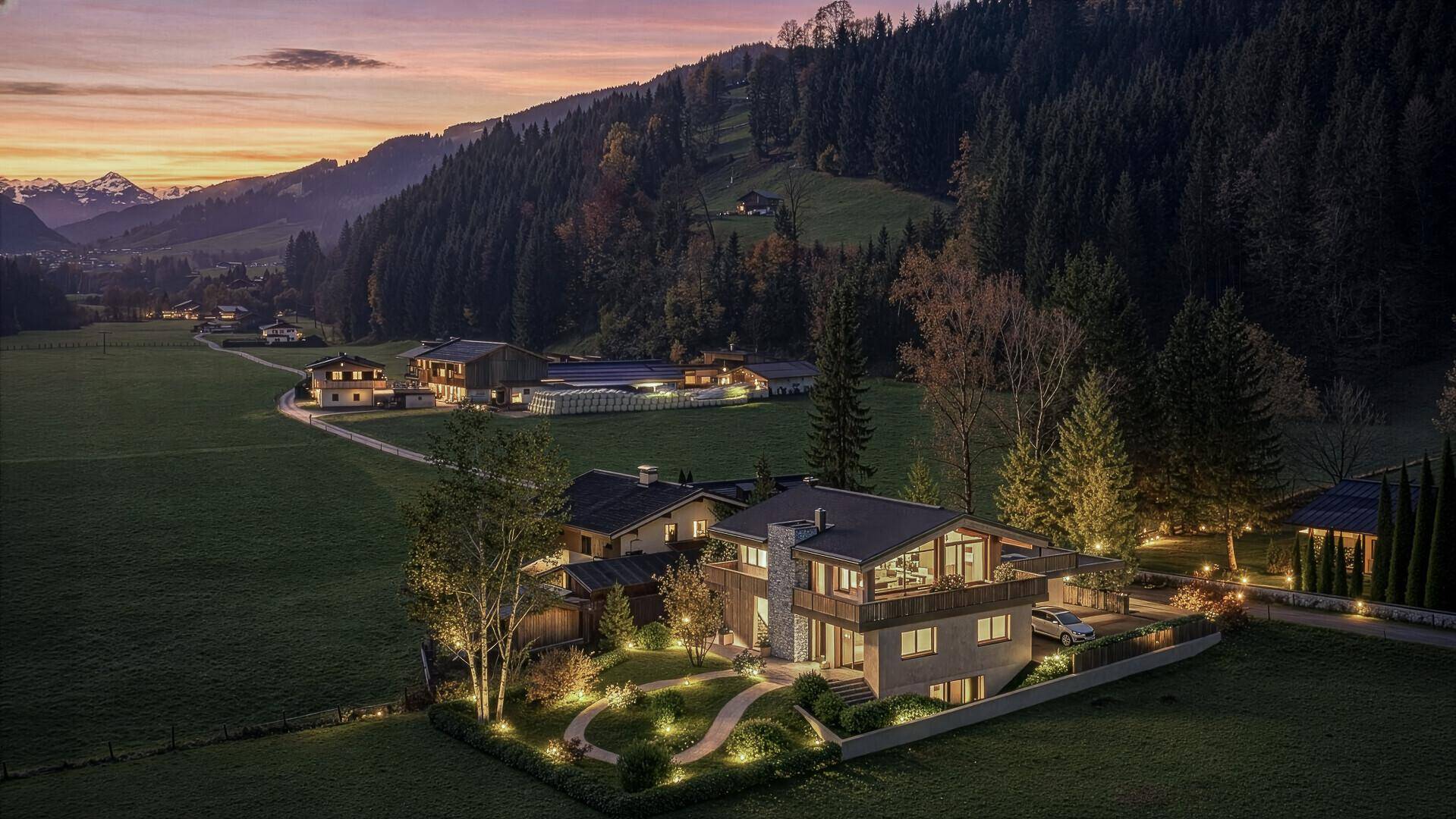 Abendliche Ansicht des Anwesens mit beleuchtetem Garten und der umliegenden Berglandschaft bei Sonnenuntergang.