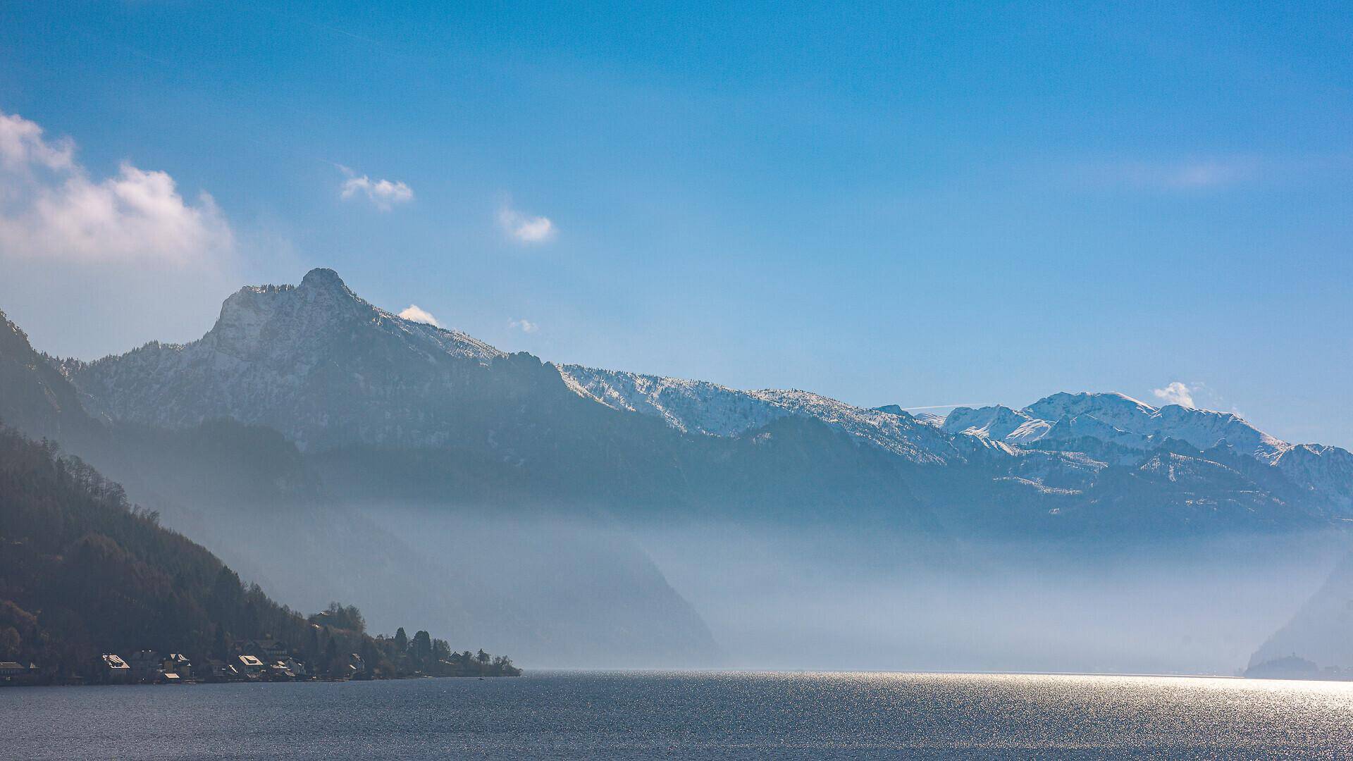Panoramablick über den See und die umliegenden Berge, teilweise in Nebel gehüllt.