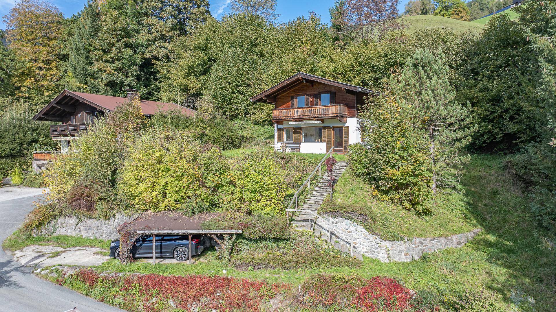 Alpines Holzhaus mit Carport und Treppe, umgeben von grünen Hügeln und Bäumen.