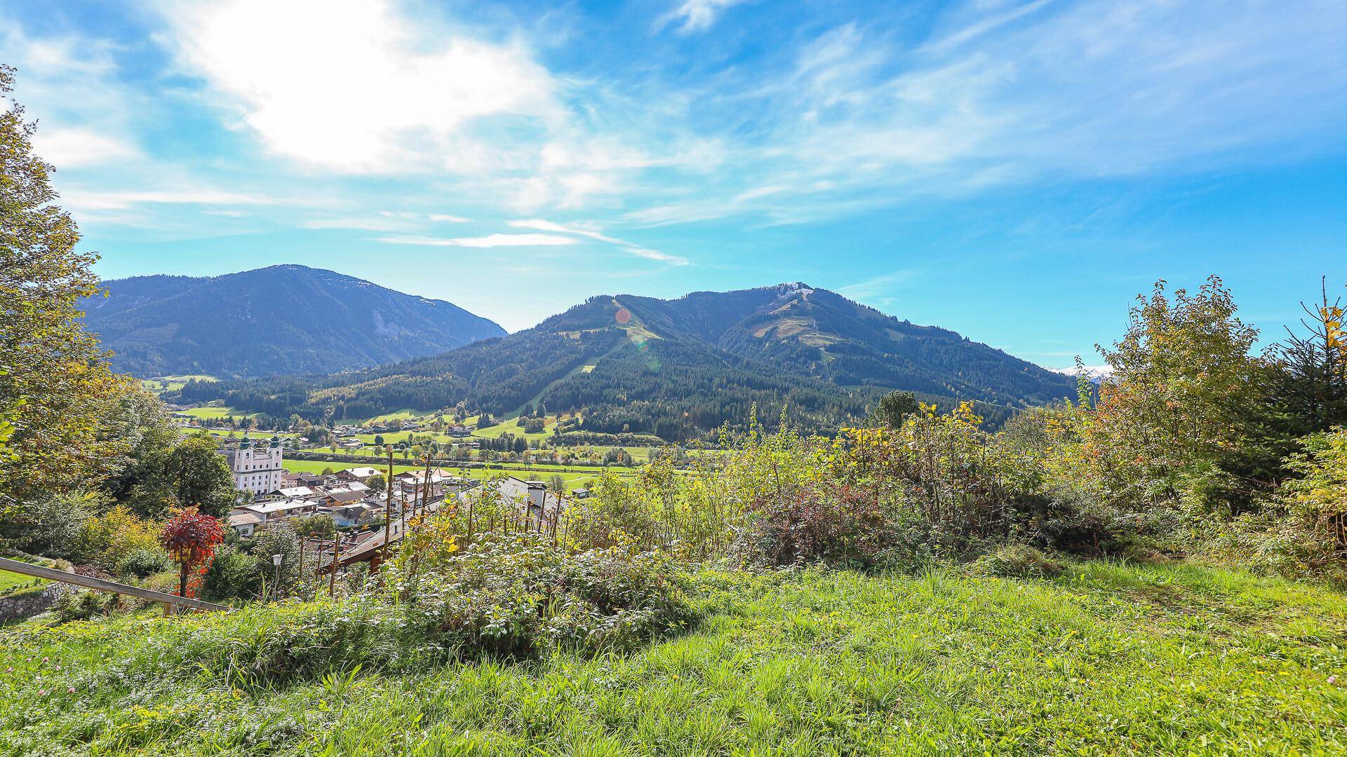 Weitläufiger Blick über grüne Hügel und ein Tal mit einem Dorf unter blauem Himmel.