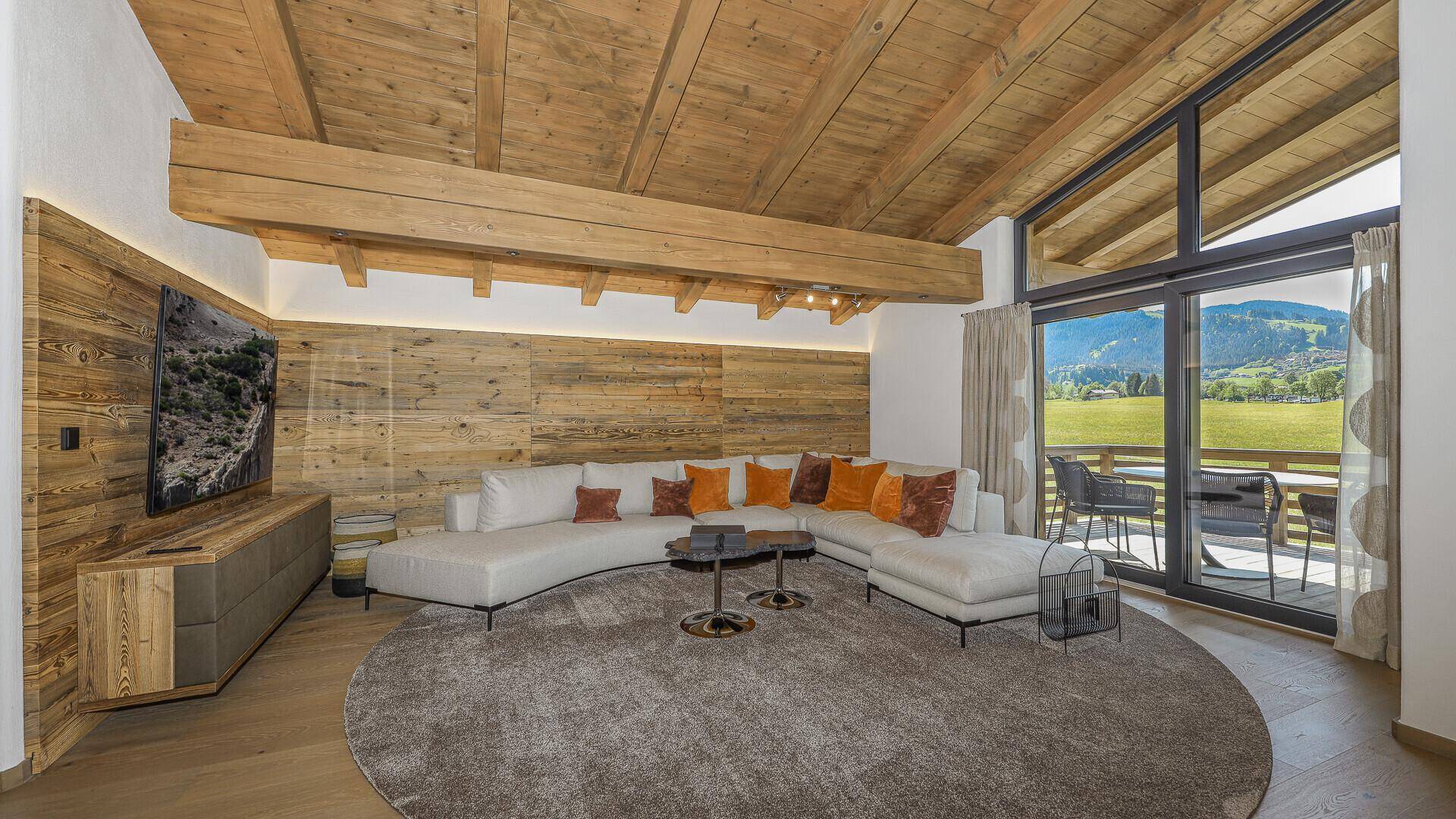 Geräumiges Wohnzimmer mit bequemer Couch, Holzwand und Zugang zum Balkon mit Bergblick.
