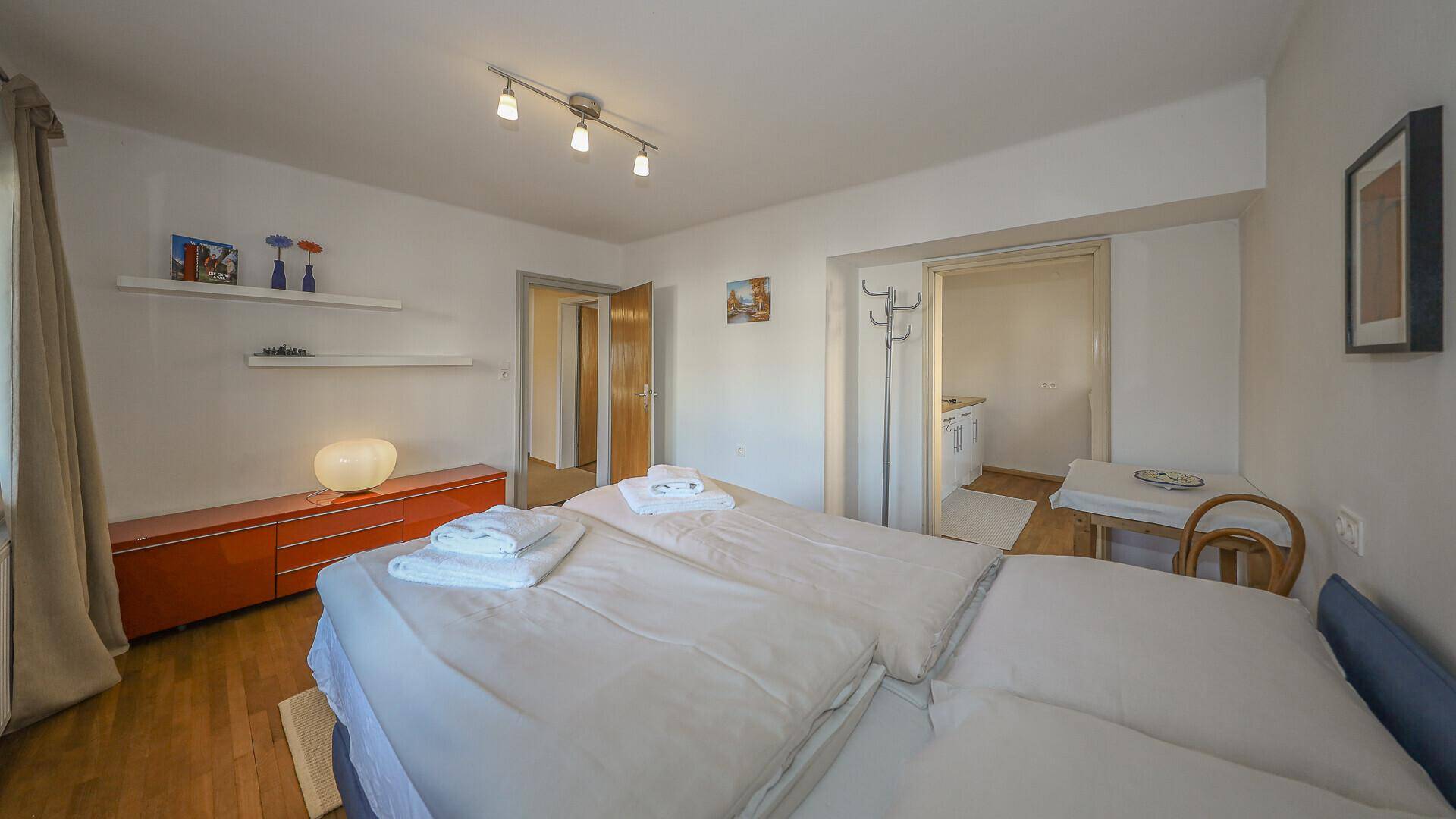 Schlafzimmer mit Doppelbett, Kommode und Zugang zu einem weiteren Raum, möglicherweise einer Küche.