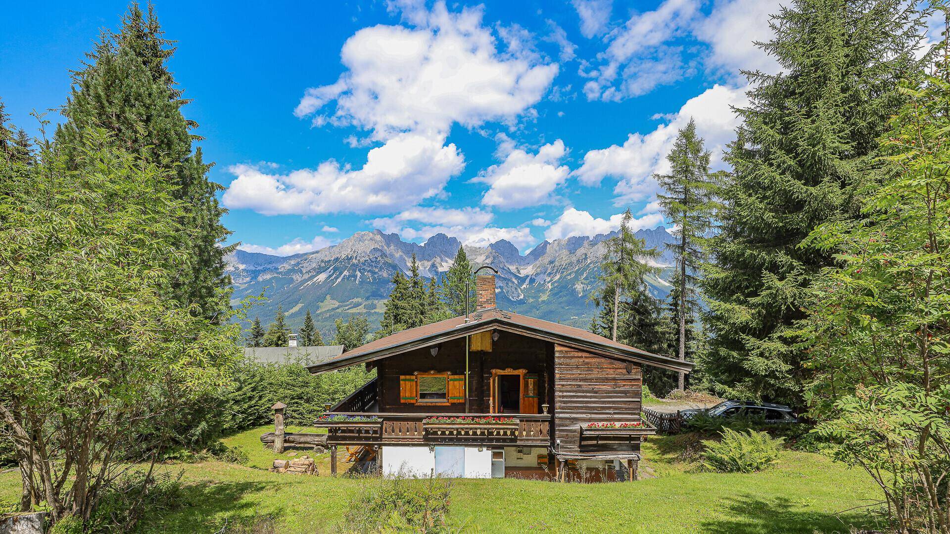 Rustikales Holzhaus mit Bergblick, umgeben von einer weitläufigen Wiese und hohen Bäumen.