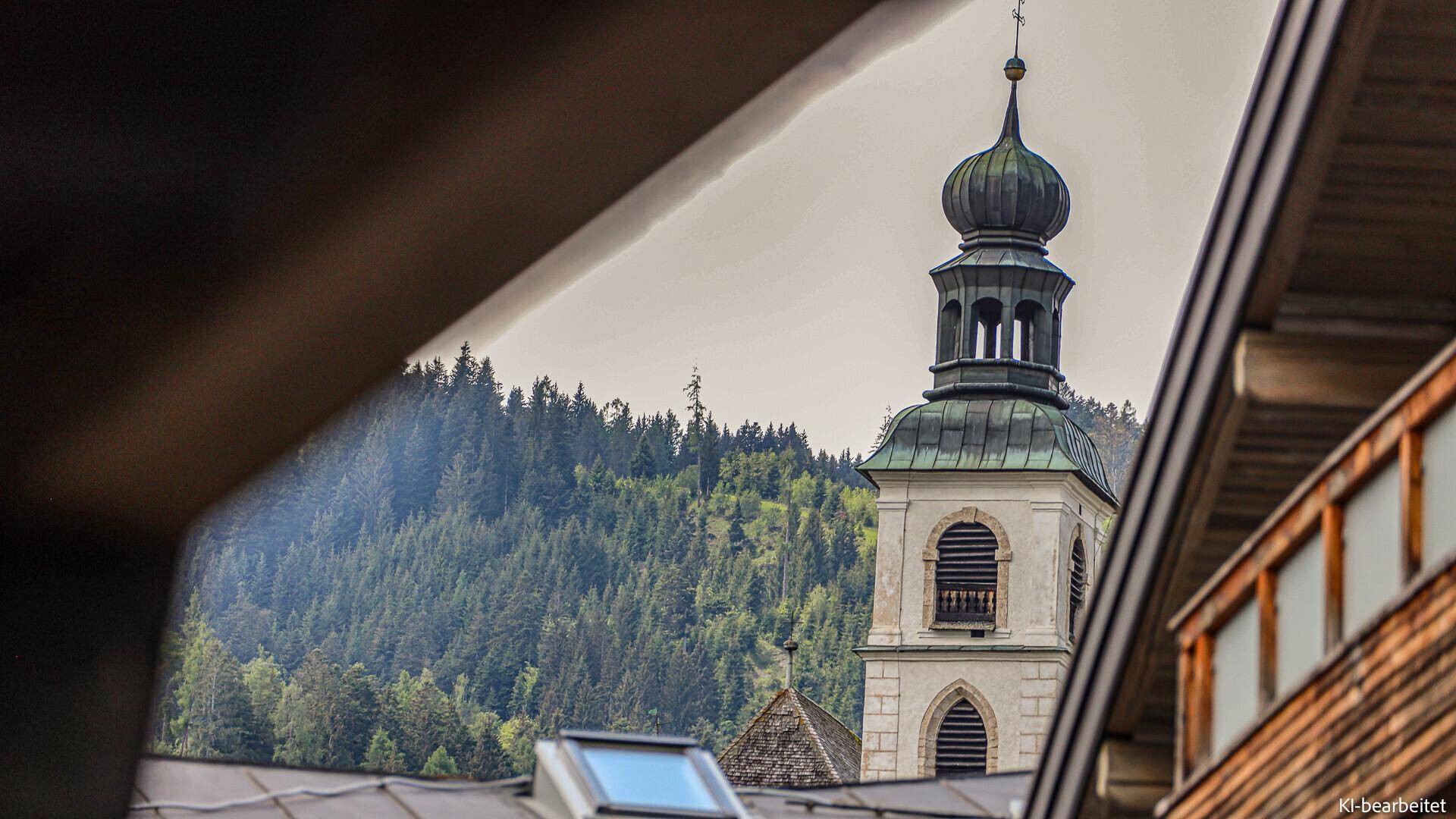 Blick auf einen Kirchturm und bewaldete Berge, eingerahmt von einem Dachgiebel.