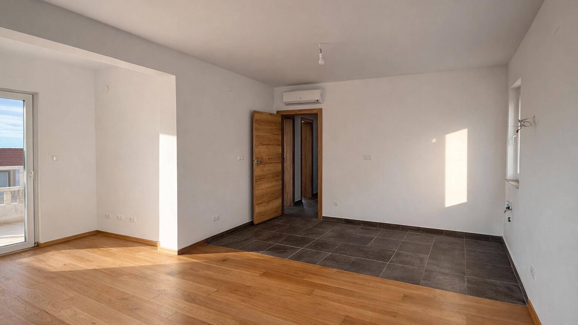 Geräumiger Raum mit gemischtem Holz- und Fliesenboden, Klimaanlage und Zugang zu weiteren Zimmern.