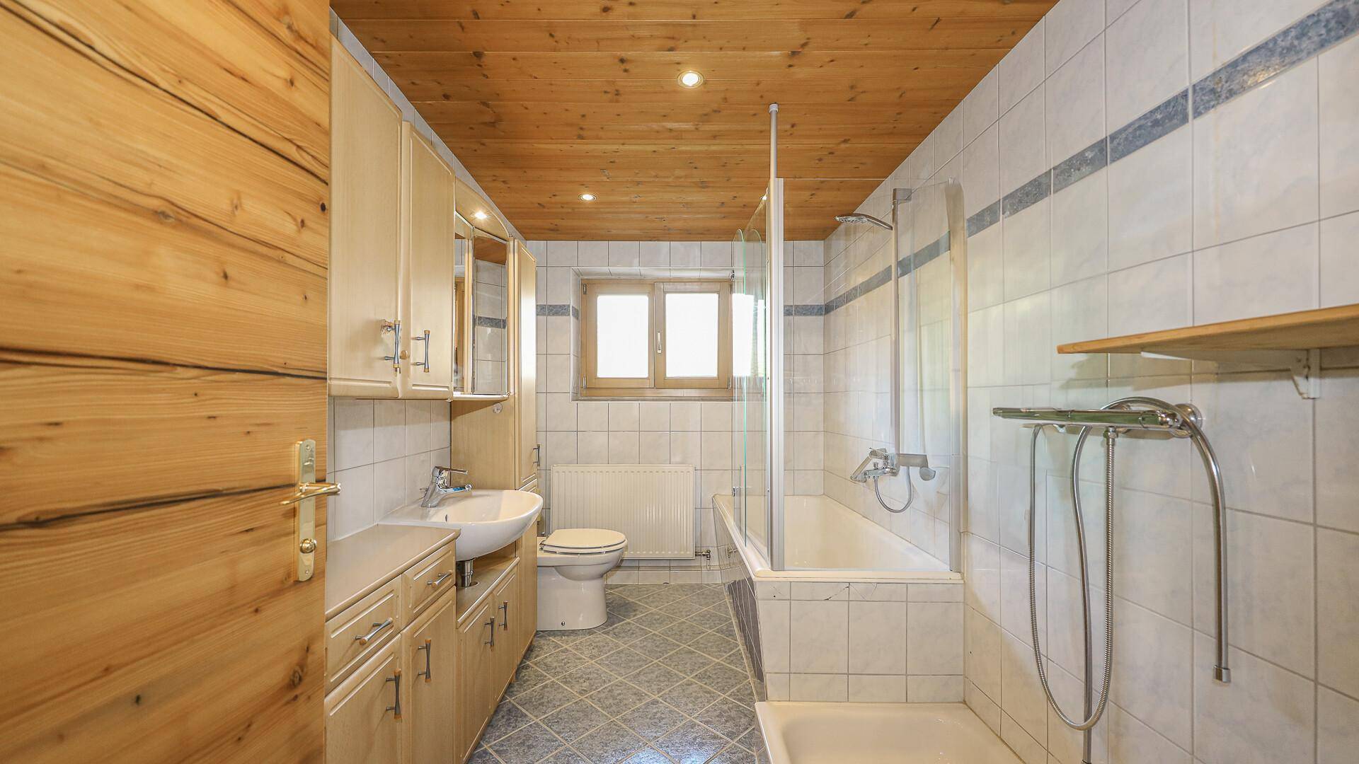 Badezimmer mit Badewanne, Dusche und Fenster, ausgestattet mit hellen Fliesen und Holzdecke.