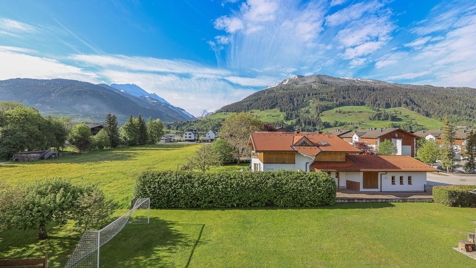 Weitläufiger Gartenbereich mit gepflegter Hecke und einem Fußballtor, umgeben von grünen Bergen.