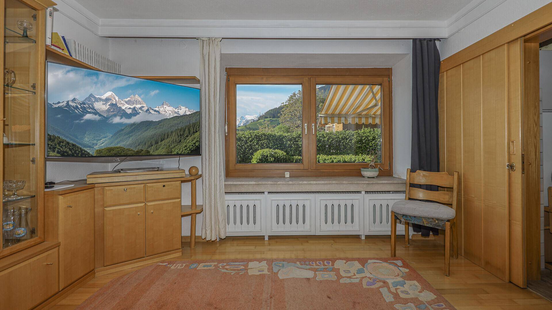 Gemütliches Wohnzimmer mit Holzmöbeln, großem Fenster und Blick auf die grüne Landschaft.
