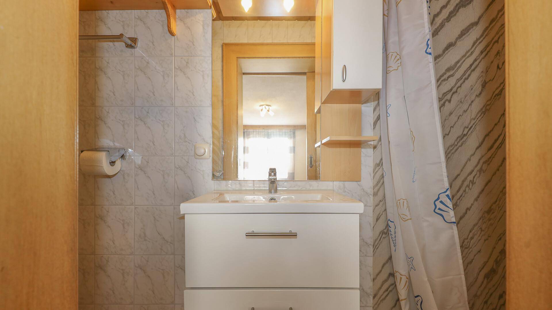 Kleines Badezimmer mit Waschbecken, Spiegel und Dusche, hell gefliest und praktisch.