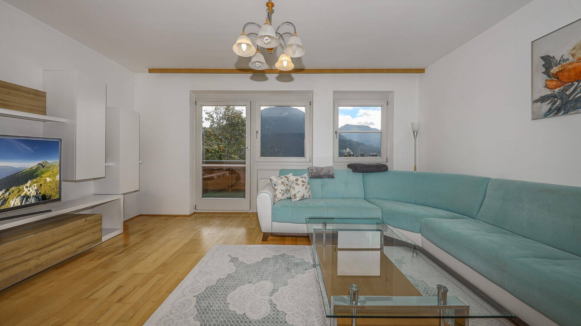 Helles Wohnzimmer mit türkisem Ecksofa, Holzboden und Bergblick durch die Fenster.