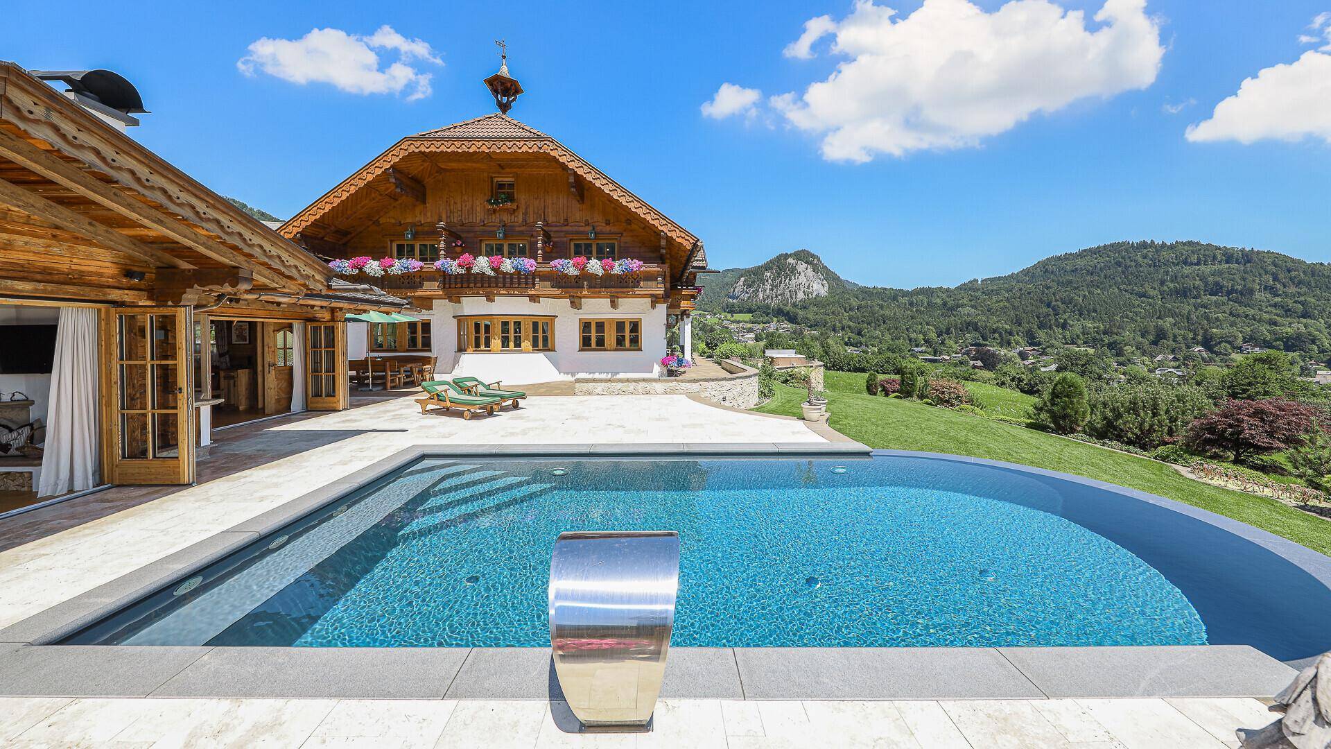 Außenbereich mit großem Pool, Sonnenterrasse und traditionellem Haus im Hintergrund mit Bergblick.