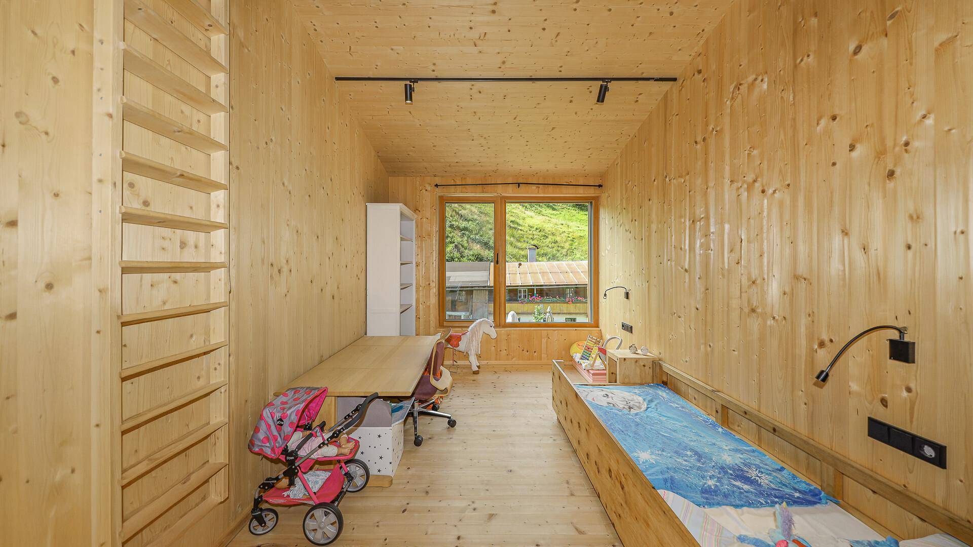 Kinderzimmer mit Holzwänden, Einzelbett, Schreibtisch und Blick auf die grüne Umgebung.