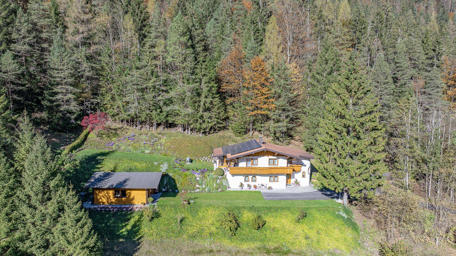 Weitläufige Luftaufnahme eines Alpenchalets mit Nebengebäude, eingebettet in eine bewaldete Berglandschaft.