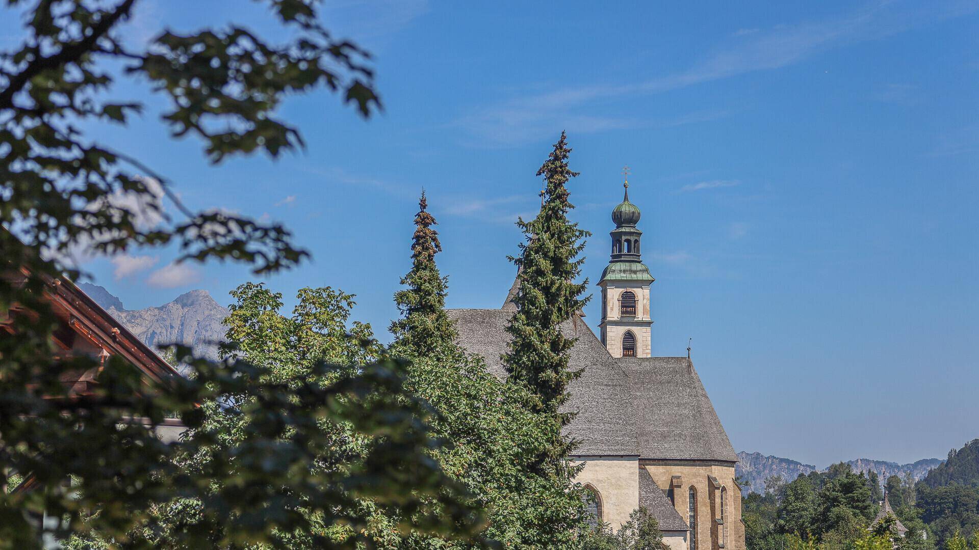 Historische Kirche mit Glockenturm, umgeben von Bäumen und Bergen im Sonnenschein.