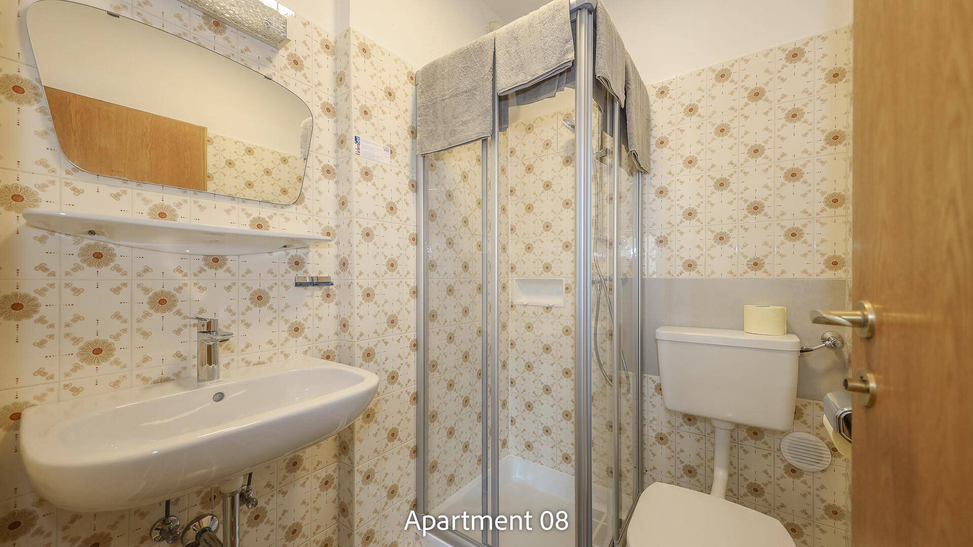 Badezimmer von Apartment 08 mit gemusterten Fliesen, Waschbecken, WC und einer Duschkabine.