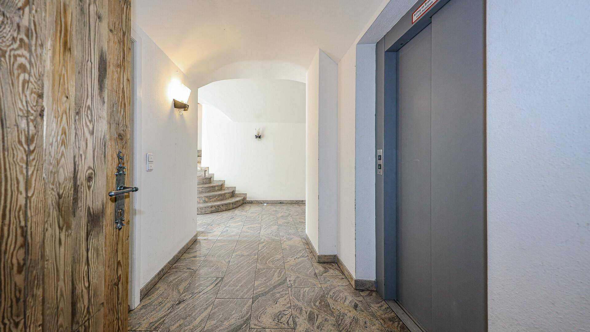Eingangsbereich mit Aufzug, Steinfliesenboden und einer geschwungenen Treppe im Hintergrund.
