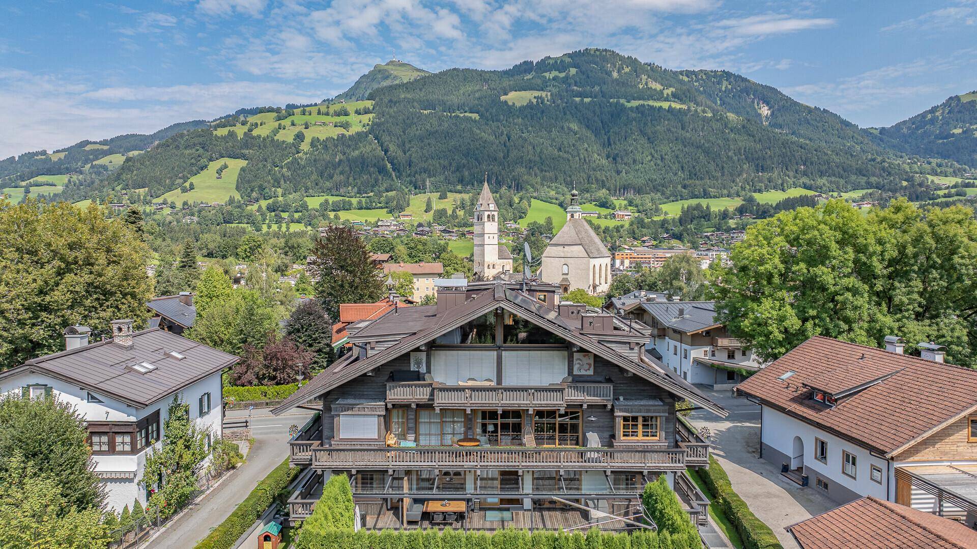 Panoramablick auf ein luxuriöses Gebäude in den Alpen, umgeben von grünen Bergen und traditionellen Häusern.