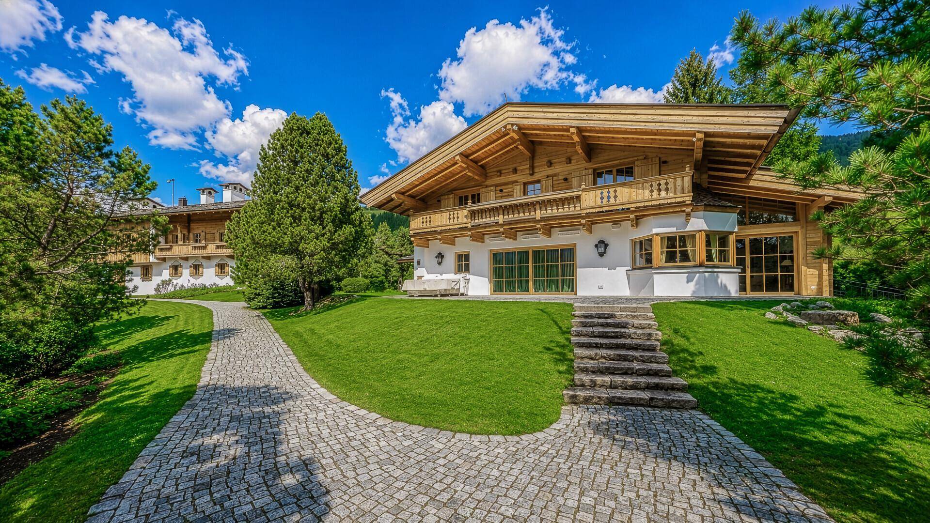 Großzügiges Anwesen im alpinen Stil mit gepflasterter Auffahrt und gepflegtem Gartenbereich unter blauem Himmel.