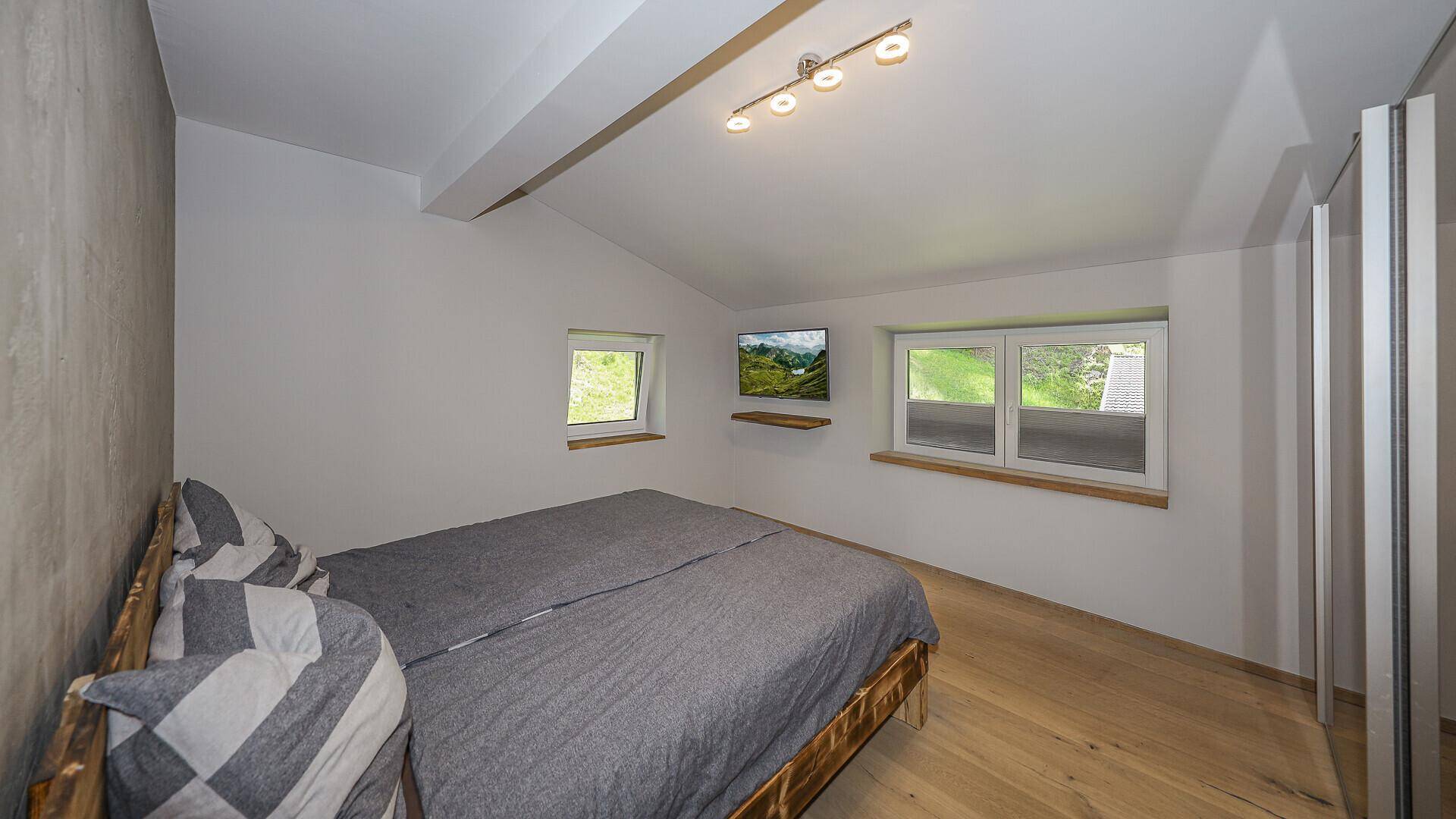 Helles Schlafzimmer mit Holzbett, Parkettboden und Fenstern mit Blick ins Grüne.