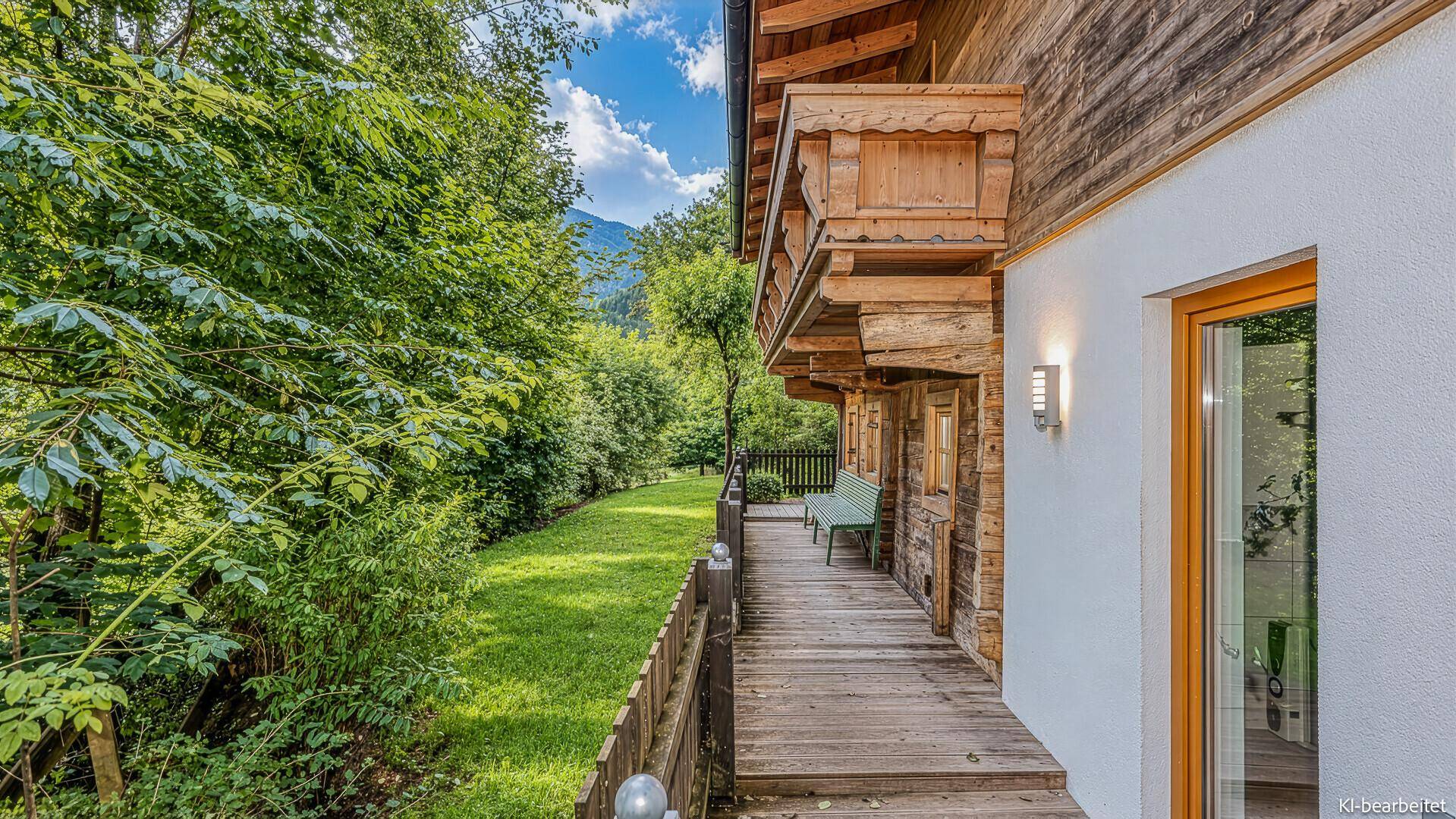 Seitliche Holzterrasse entlang des Hauses, umgeben von üppigem Grün und Bäumen.