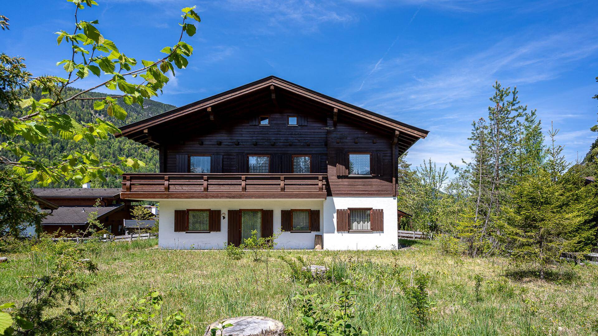 Frontansicht eines traditionellen Chalets mit Holzbalkon und weißer Fassade vor alpiner Kulisse.