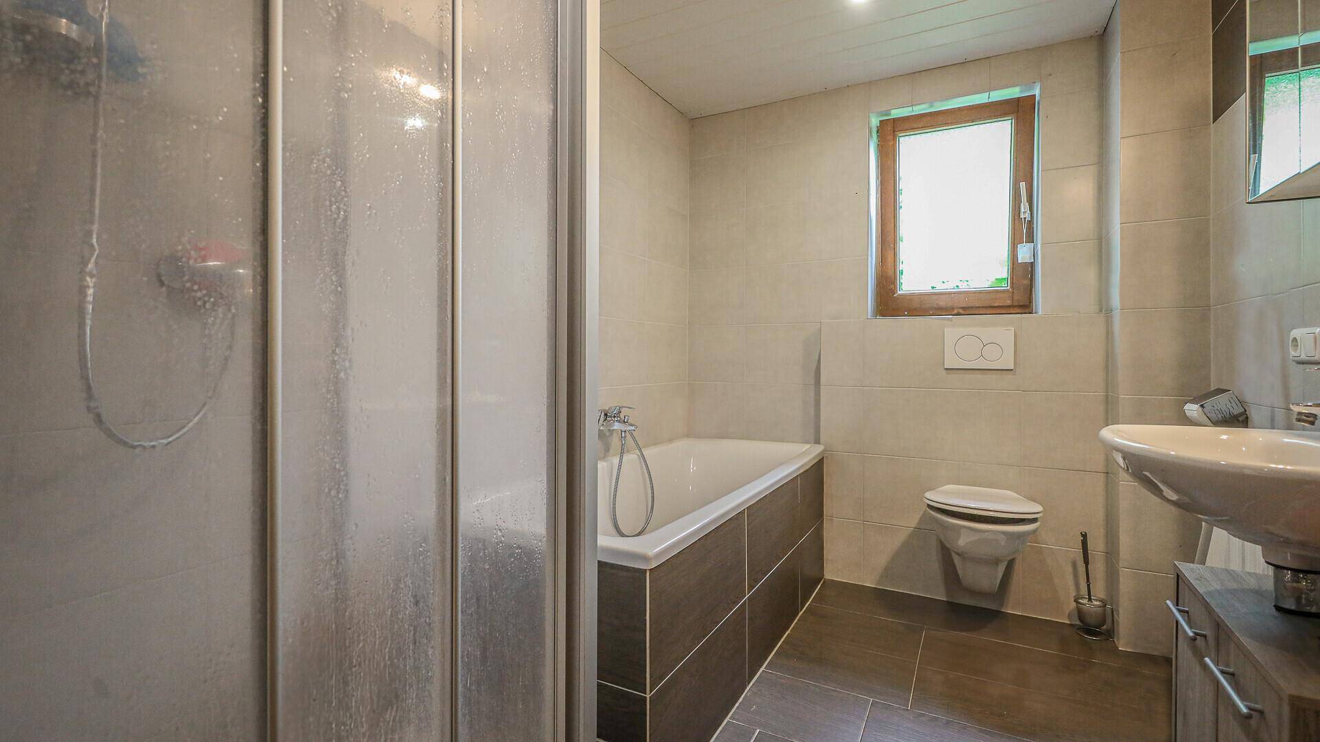 Helles Badezimmer mit Badewanne, Dusche und Fenster, ausgestattet mit weißen Fliesen.