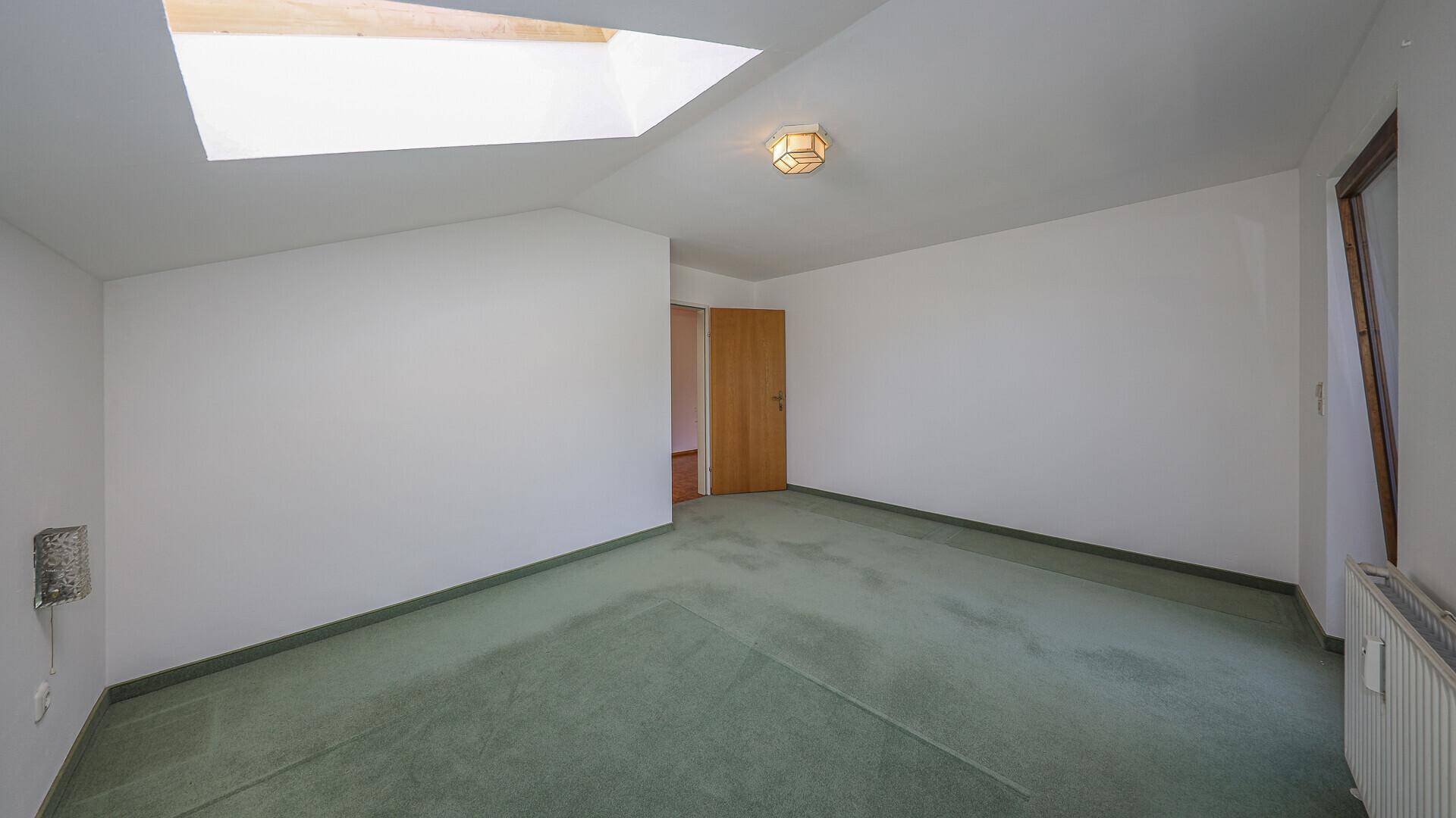 Leeres Schlafzimmer mit grünem Teppichboden, Dachfenster und Zugang zu einem weiteren Raum.