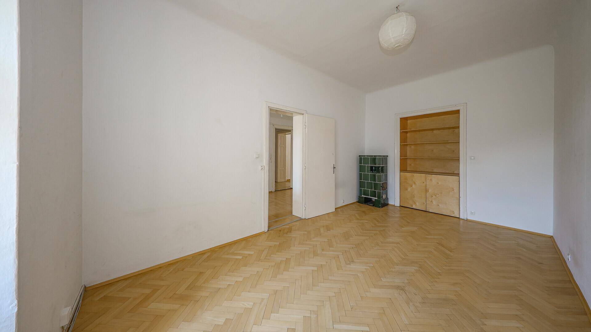 Helles Zimmer mit Parkettboden, Kachelofen und eingebauten Holzregalen.