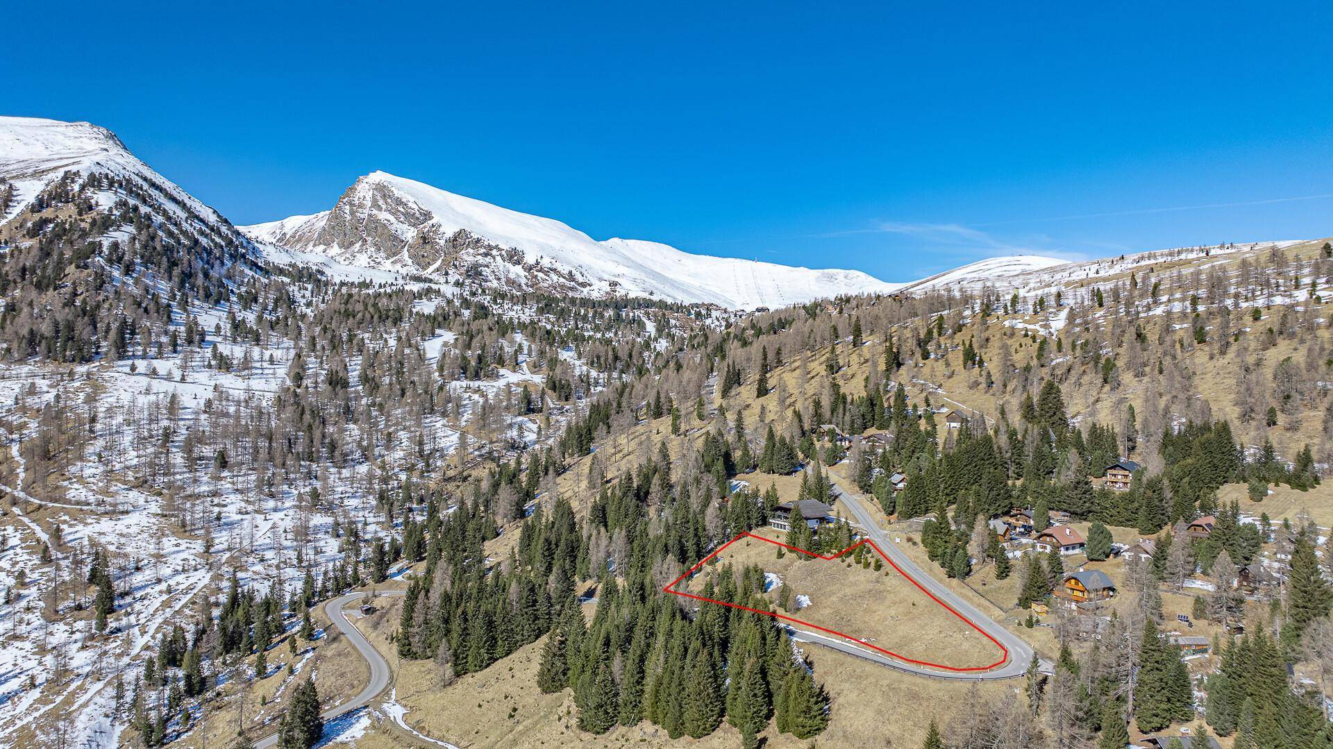 Luftaufnahme einer alpinen Landschaft mit schneebedeckten Bergen und einem markierten Grundstück unter strahlend blauem Himmel.