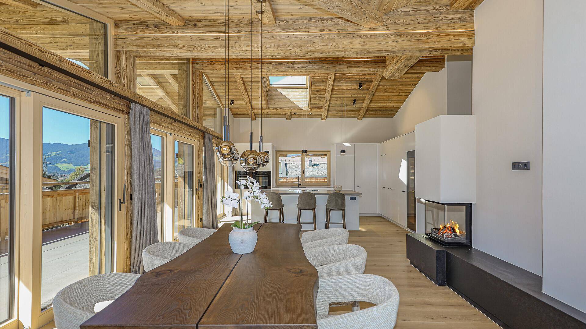 Offener Wohn- und Essbereich mit Kamin, Holzbalkendecke und moderner Küche im Hintergrund.