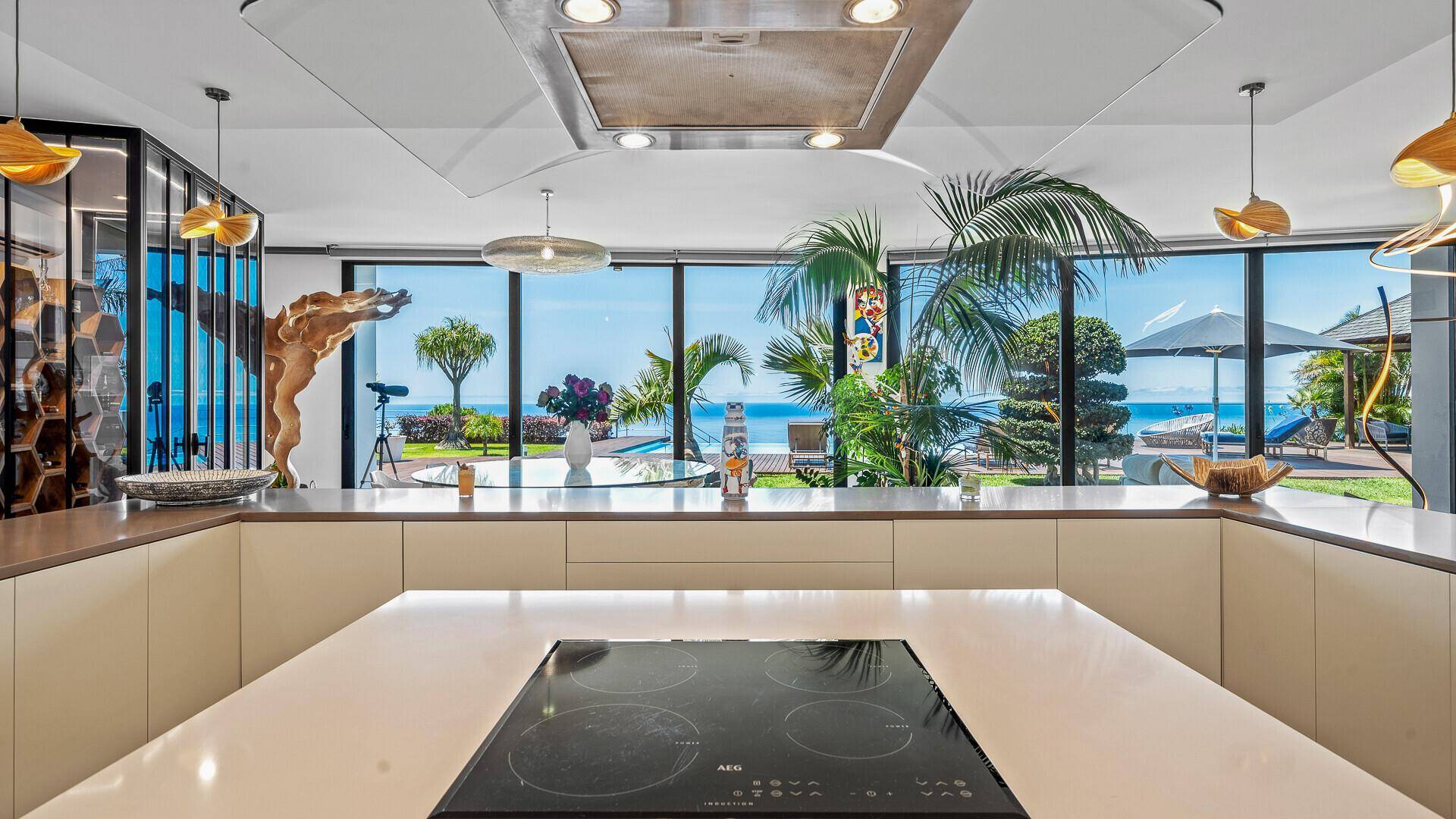 Küchenarbeitsplatte mit Induktionskochfeld und weitem Blick durch große Fenster auf den Garten, Pool und das Meer.