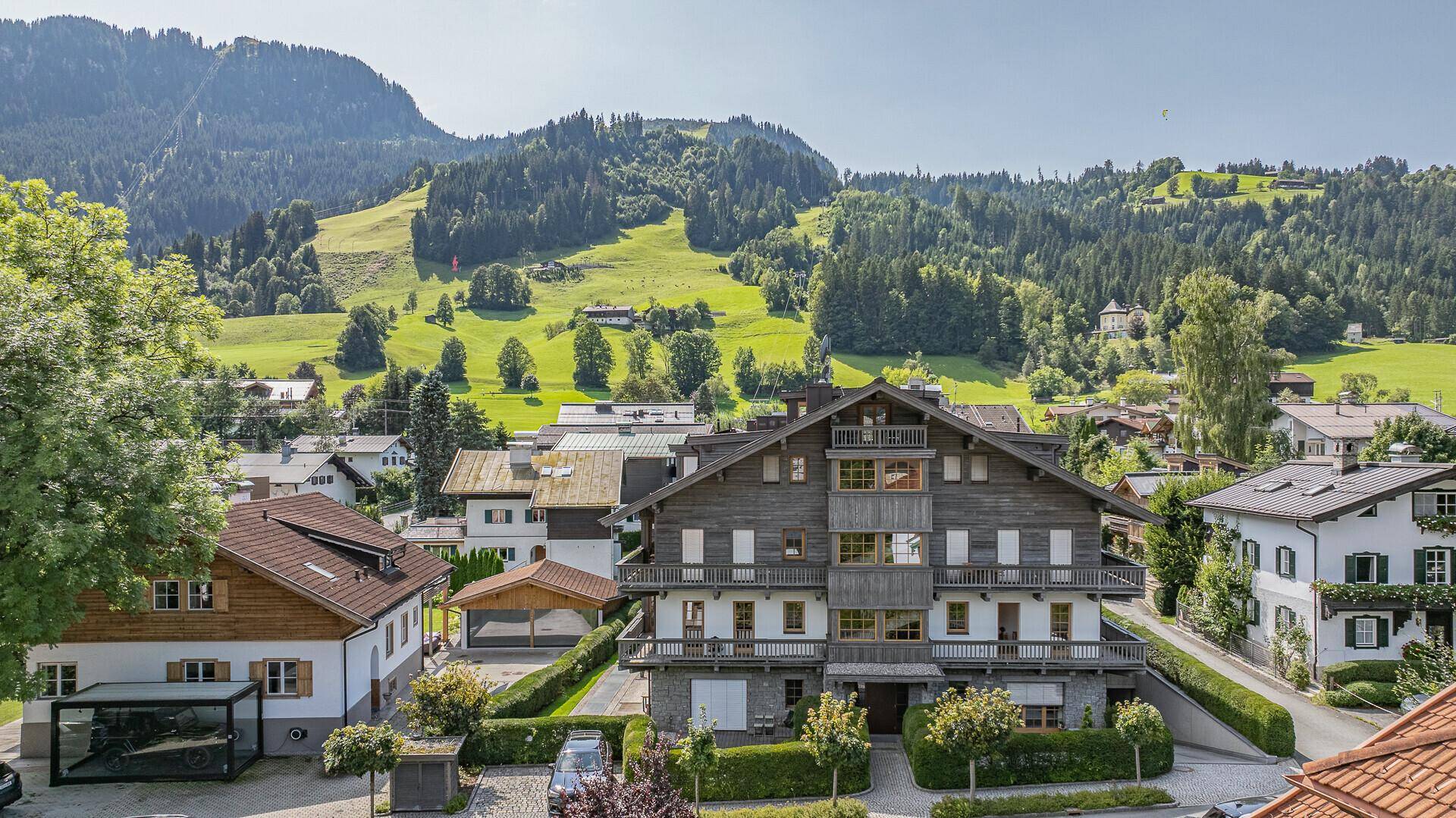 Traditionelles Gebäude mit Holzbalkonen vor einer malerischen Bergkulisse und grünen Wiesen.