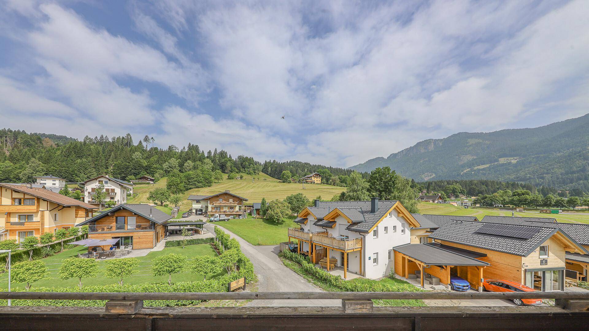 Blick von einem Balkon auf umliegende Häuser, grüne Felder und eine beeindruckende Bergkulisse.