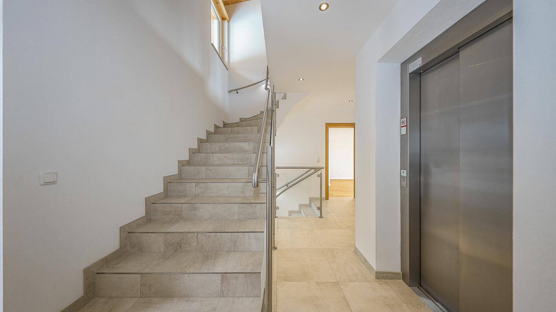 Geräumiger Eingangsbereich mit Steintreppe, Glasgeländer und modernem Aufzug.