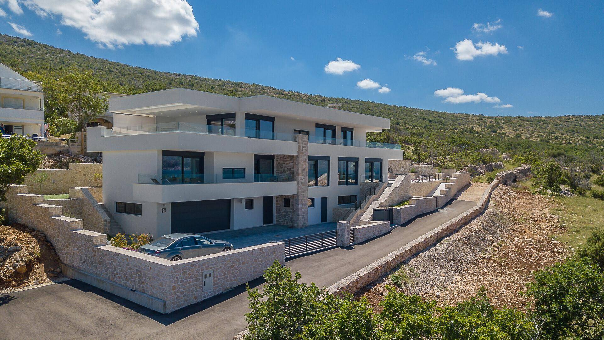 Moderne Villa mit mehreren Balkonen, großen Glasfronten und Zufahrt in einer grünen Hügellandschaft.