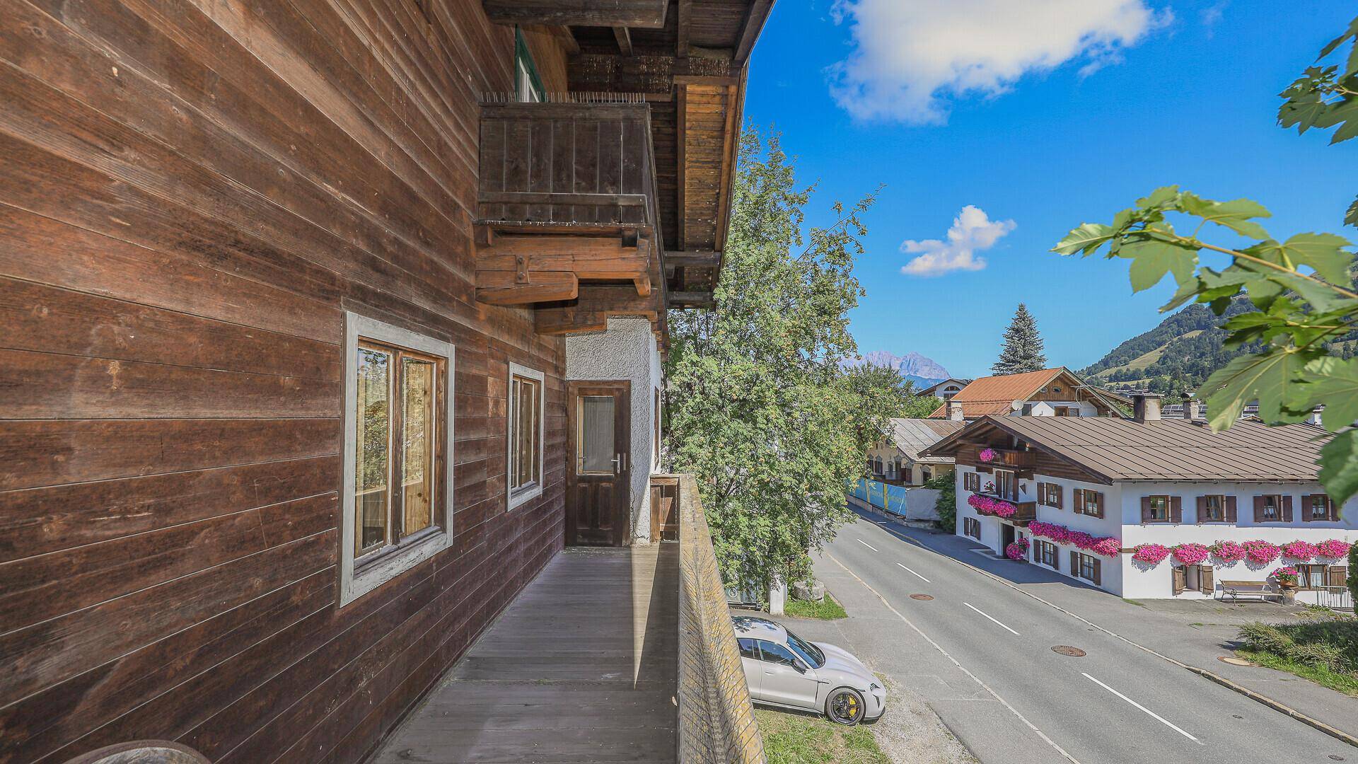 Blick vom Holzbalkon auf die Straße und die umliegenden Häuser mit Bergpanorama im Hintergrund.