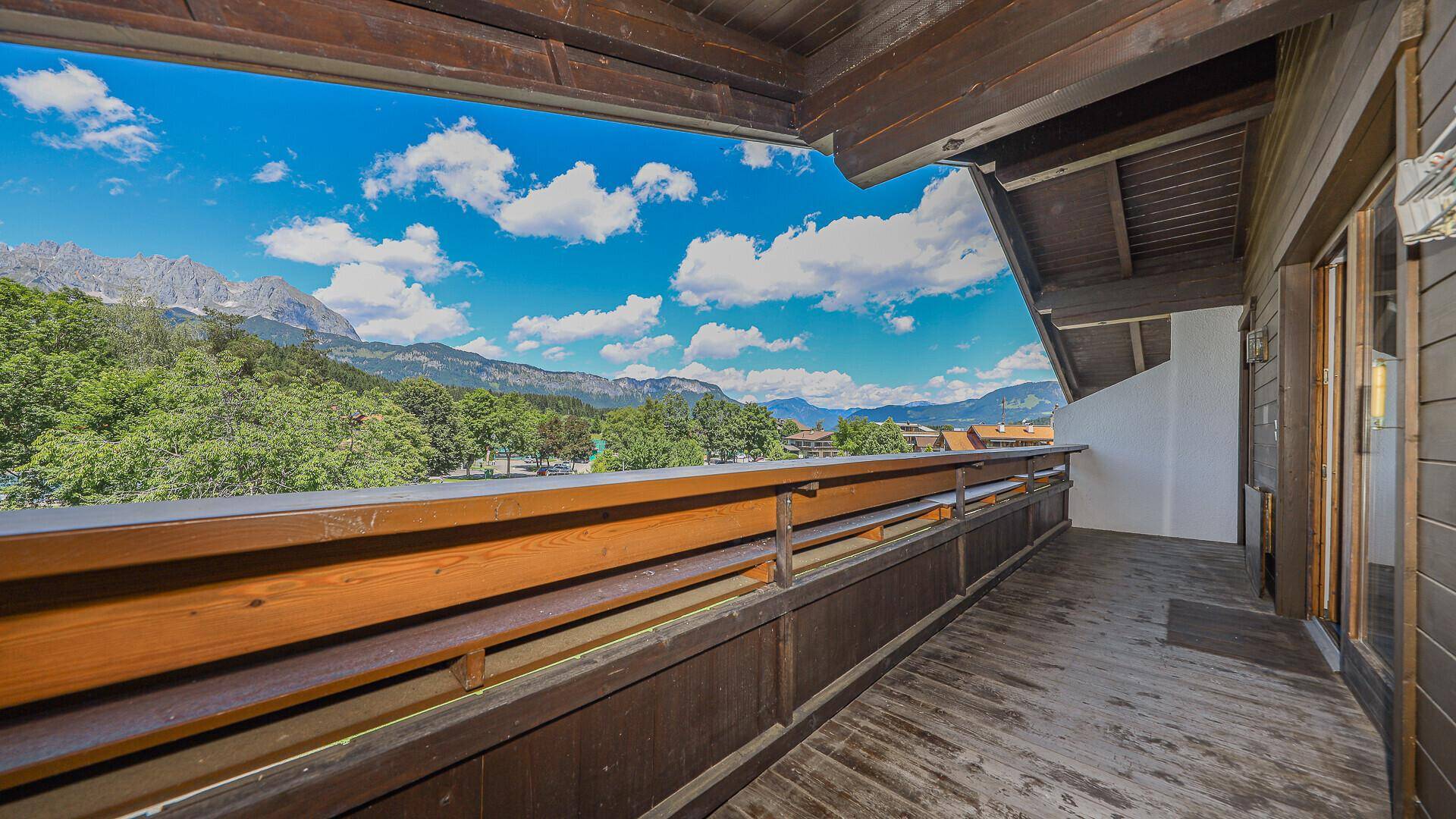 Langer Holzbalkon mit weitem Blick auf die Berge und die grüne Landschaft unter blauem Himmel.