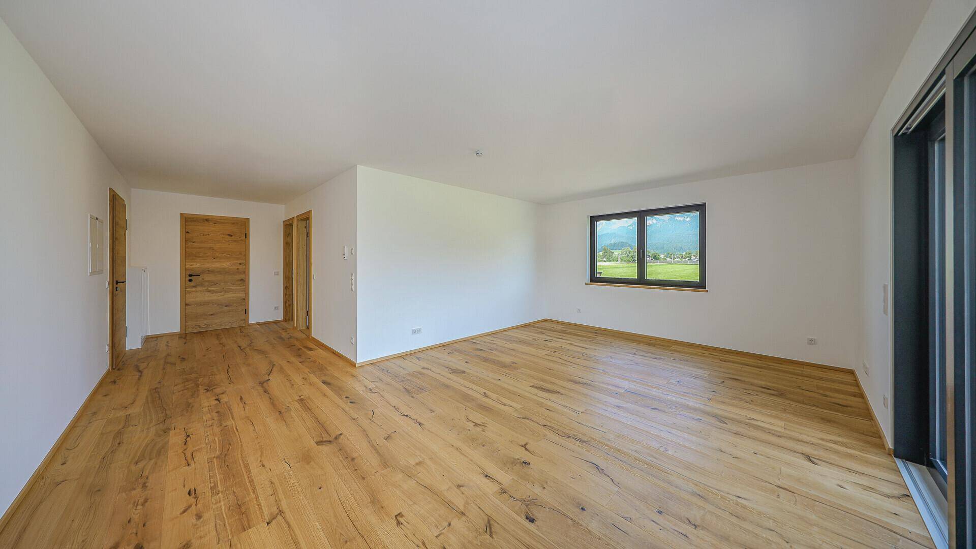 Heller, unmöblierter Raum mit Holzboden und Fenster mit Blick ins Grüne.